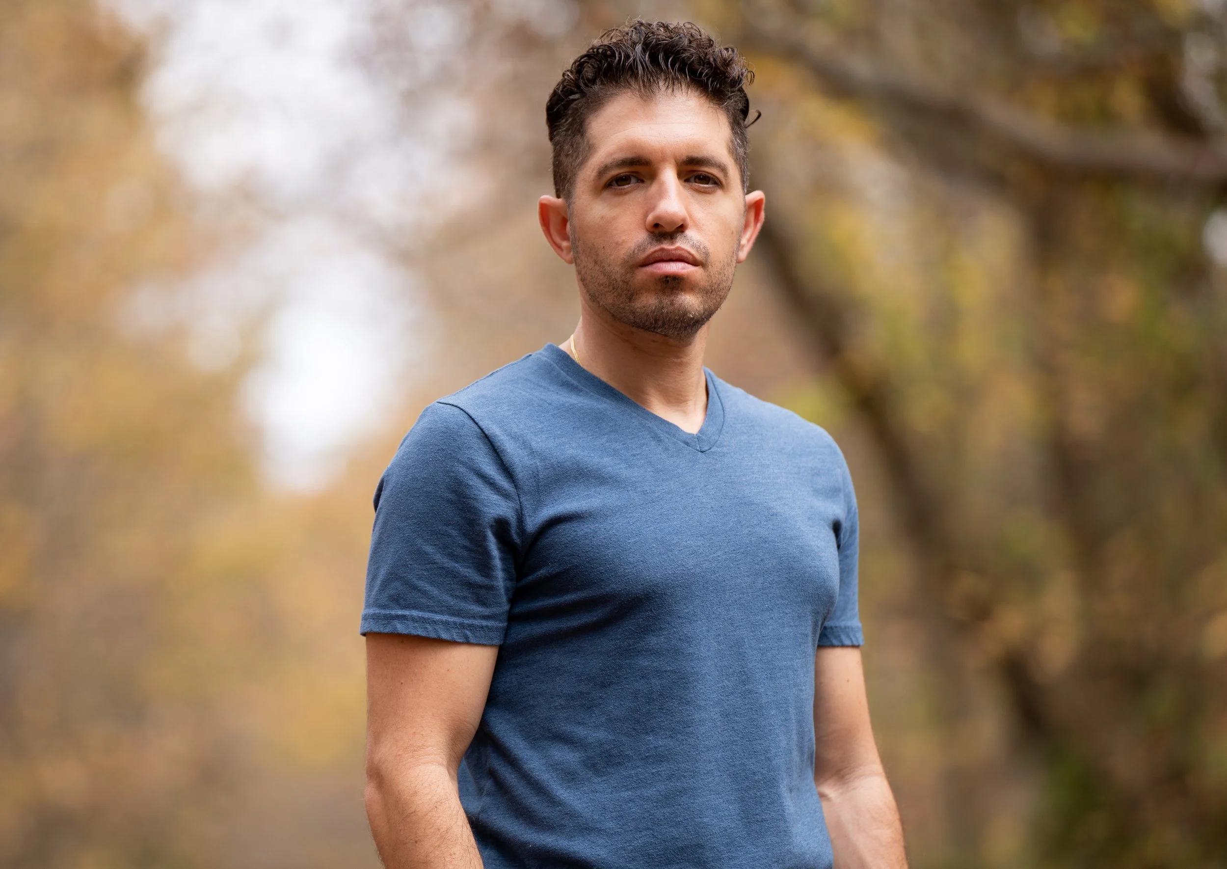 A man with short dark hair and a beard stands outdoors in front of a blurred background of trees with autumn-colored leaves, wearing a blue t-shirt.
