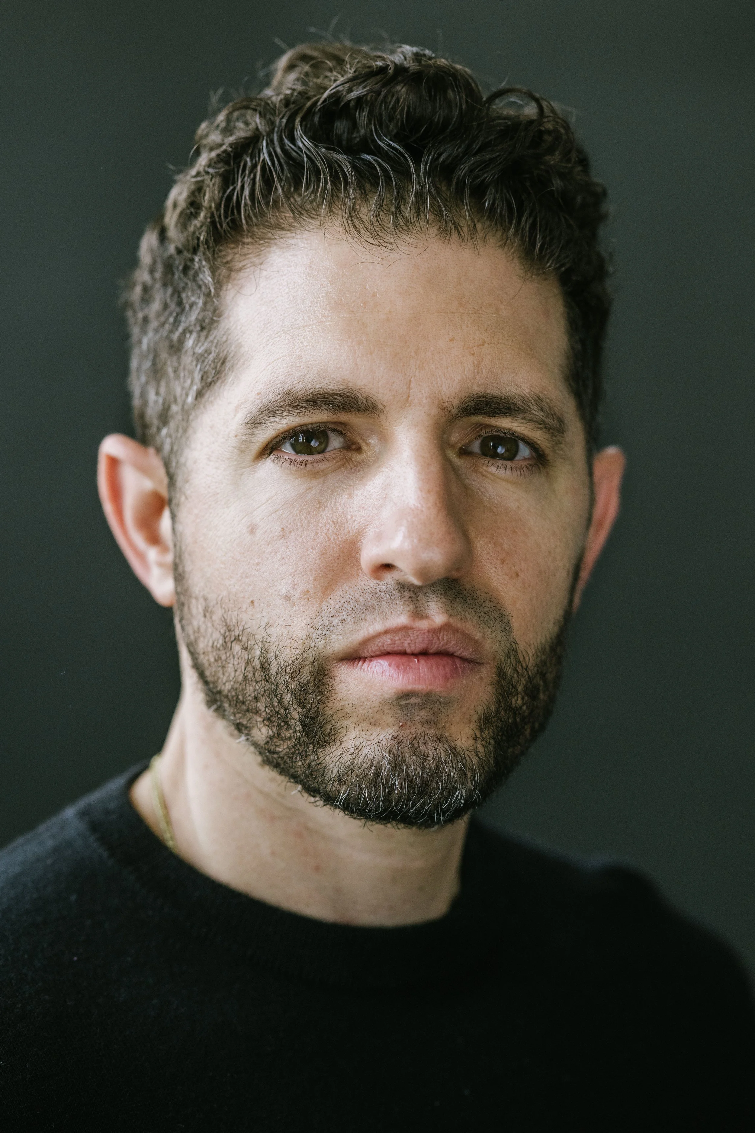 Close-up portrait of a man with dark, curly hair, a beard, and wearing a black shirt against a dark background.