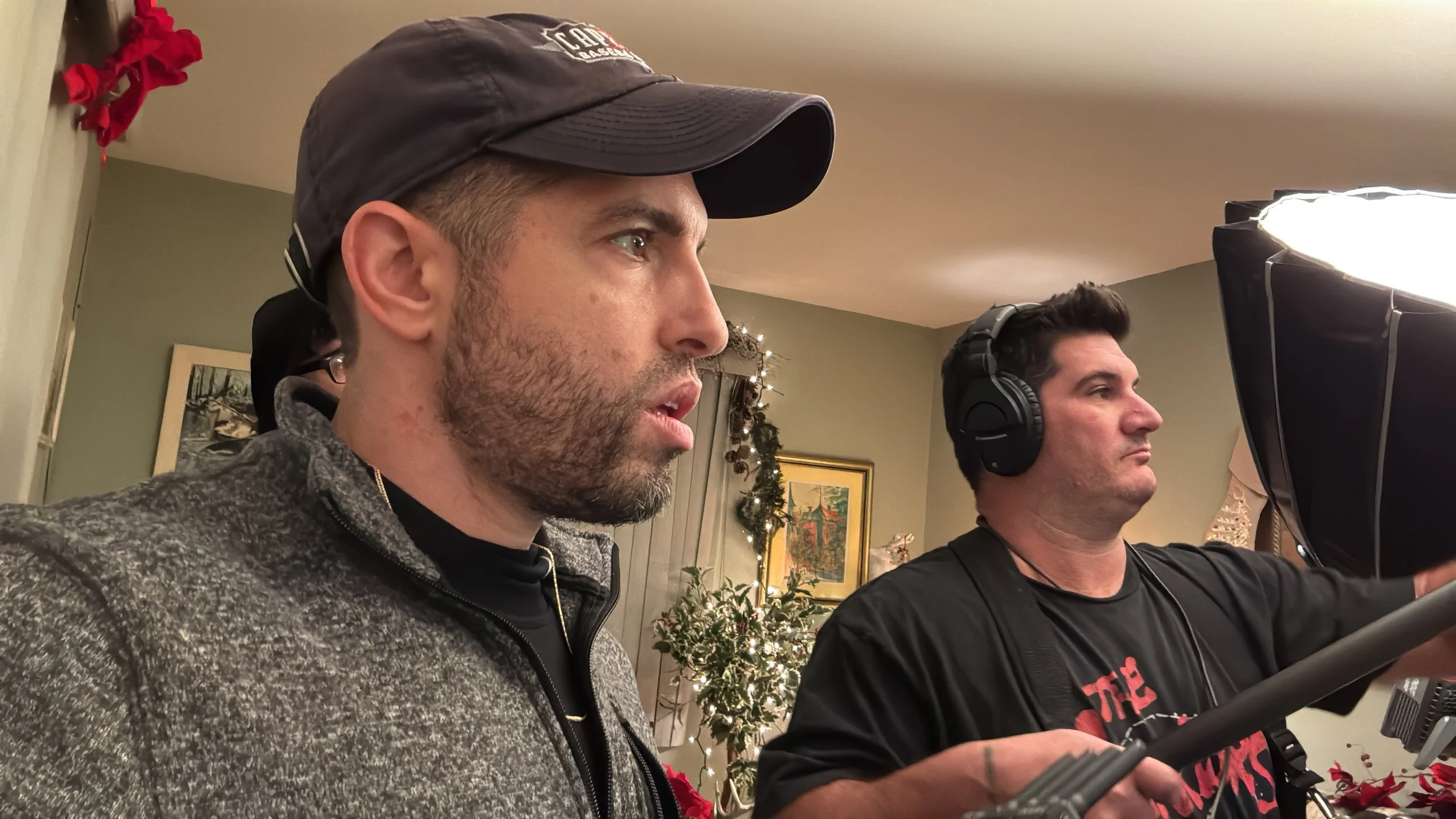 Two men on a film set, one is directing while the other is acting. The director is wearing headphones and holding a remote, focusing intently. The actor is wearing a baseball cap and a fleece jacket, concentrating on the scene. Holiday decorations are visible in the background, including a Christmas tree and red poinsettias.