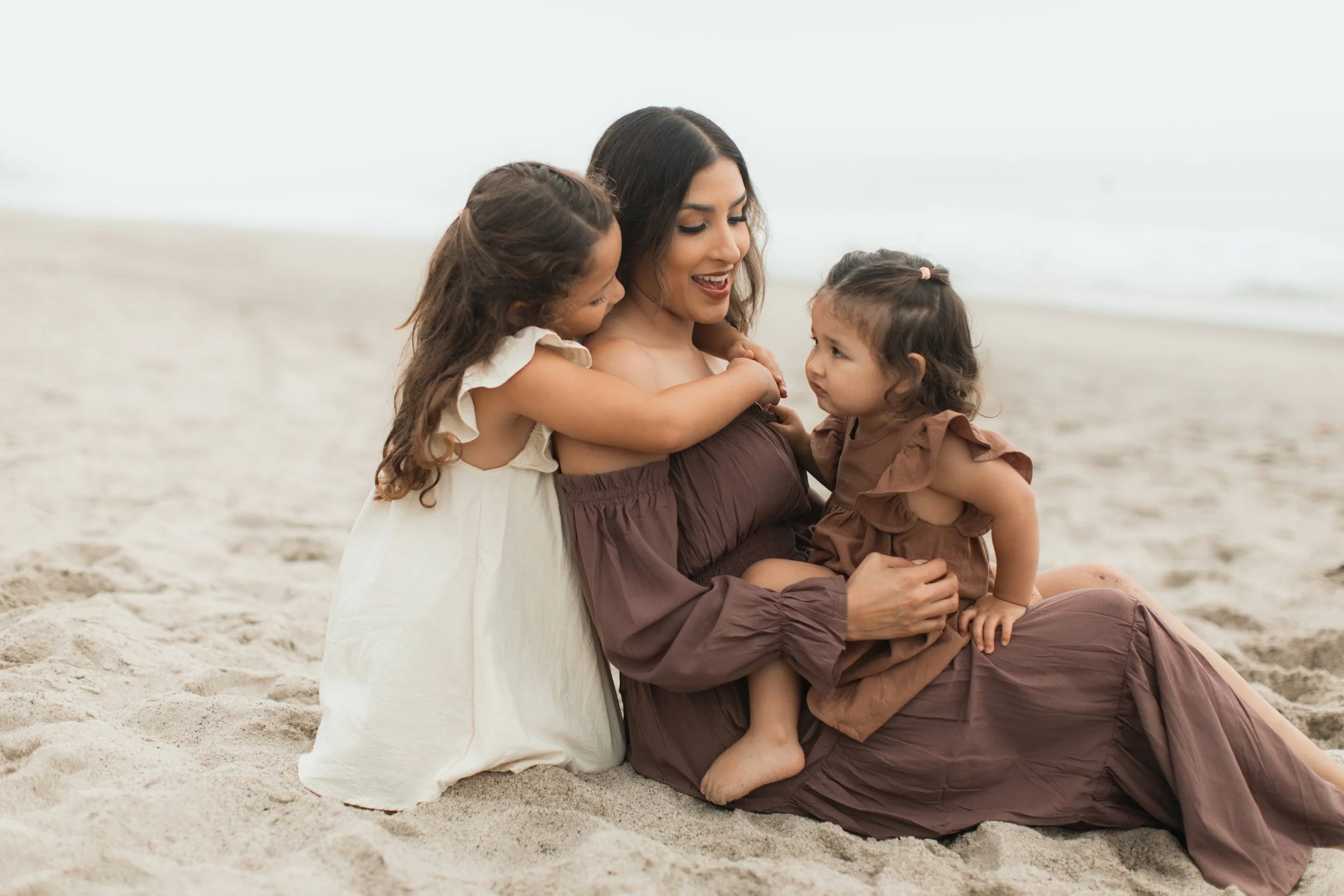 Mother sitting on a beach with two young daughters, one in a white dress, the other in a brown dress, all smiling and embracing.