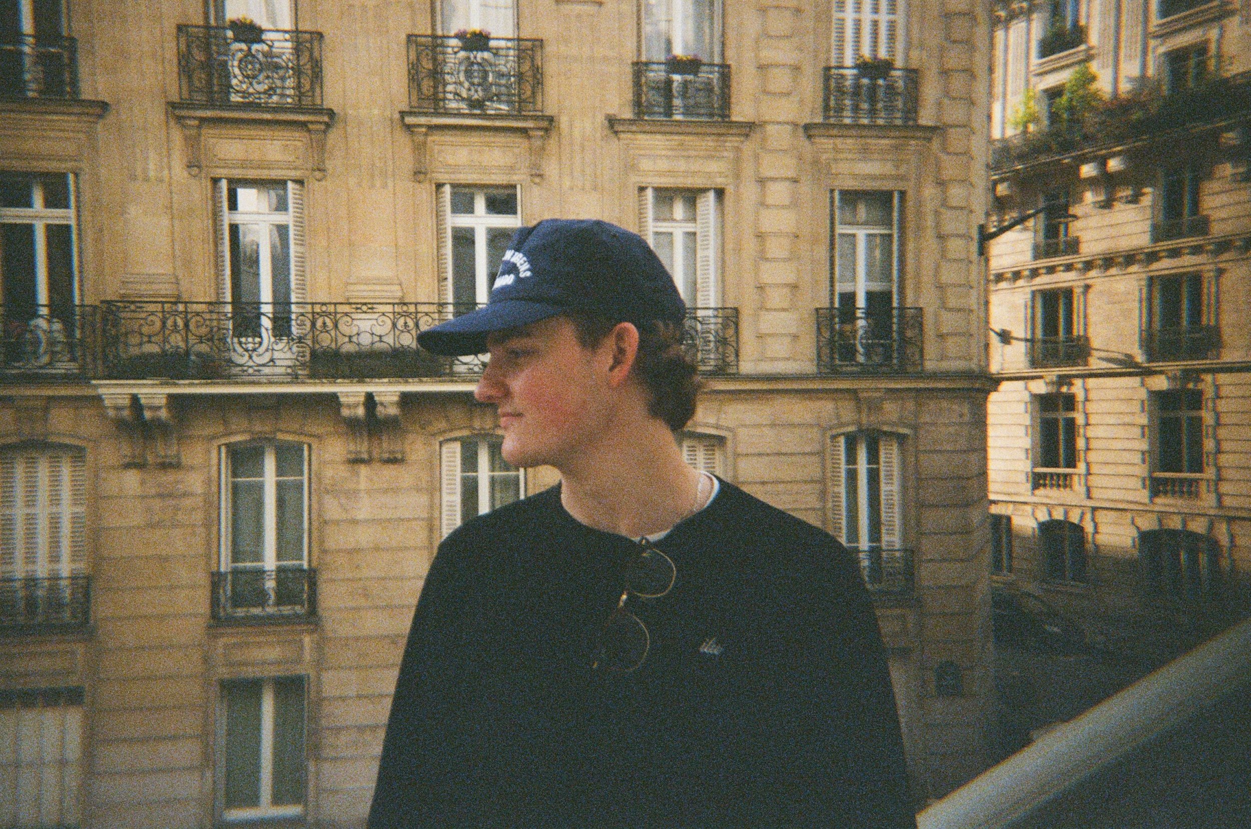A young man wearing a navy blue cap and dark sweatshirt with glasses hanging from the collar, standing on a balcony with a European cityscape background featuring classic stone buildings with large windows and wrought-iron balconies.