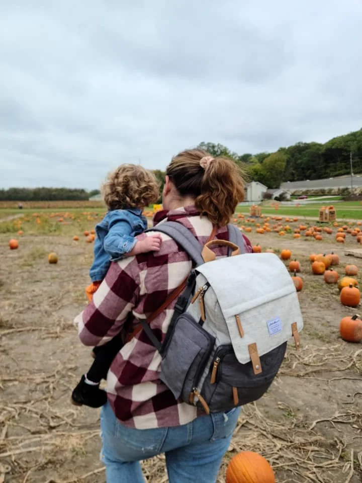 A woman with dark highlighted hair and light skin, wearing a flannel shirt and jeans, carriers a curly blonde haired toddler in a dress and a jean jacket. They are facing away from the camera and looking out over a pumpkin patch.