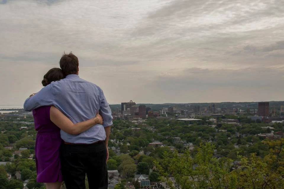 A dark haired light skinned woman in a purple dress stands with her arm around a dark haired lightly tanned man in dark slacks and a light blue dress shirt. They are facing away from the camera overlooking a town which is sprawled out below.