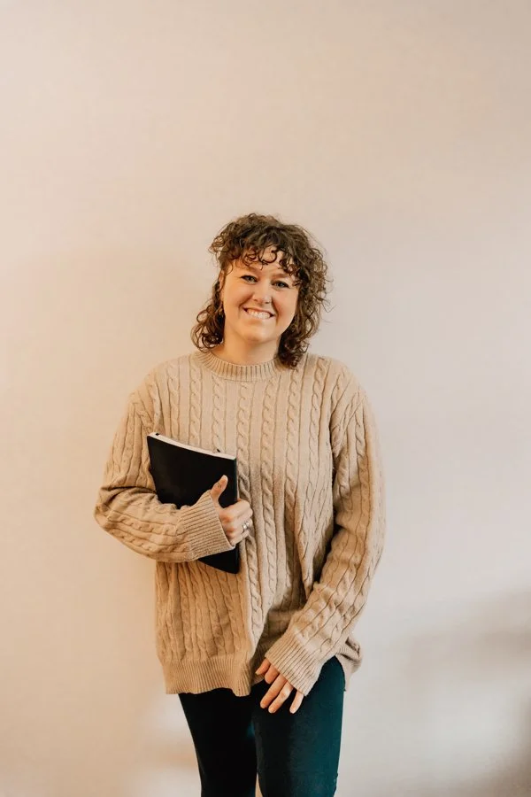 A young woman with curly hair, wearing a beige cable-knit sweater, holding a black notebook, standing against a plain white wall and smiling at the camera.
