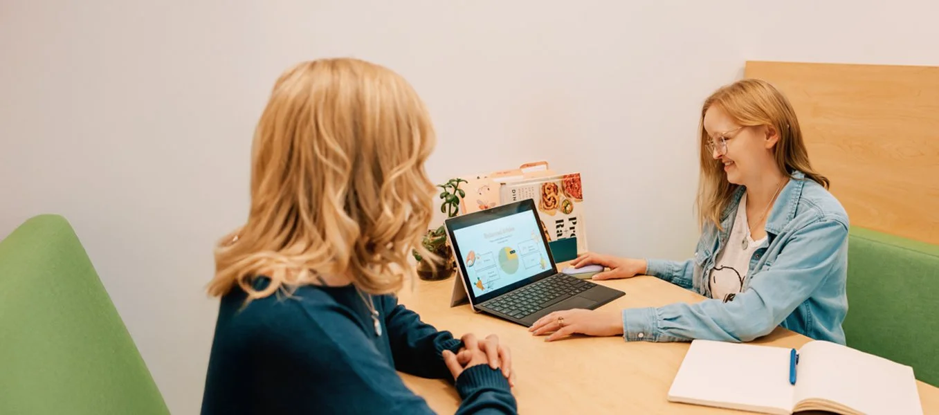 Two women sitting at a wooden table in a room with light-colored walls, engaging in a discussion. One woman, with blonde curls, is facing away, while the other woman, with straight reddish hair and glasses, is smiling. A tablet with a presentation or graphic is on the table between them.
