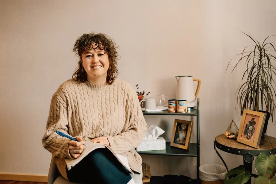 A young woman sitting on a chair, smiling, holding a pen and notebook, in a cozy room with a shelf, framed photos, and houseplants.