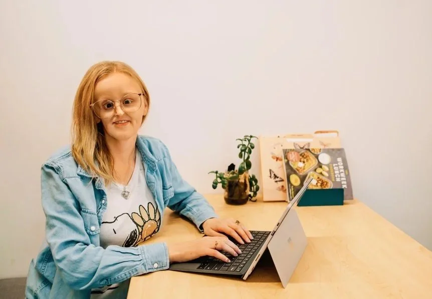 A young girl sitting at a wooden table, smiling at the camera, working on a laptop. Behind her are books, a plant, and a solid white wall.