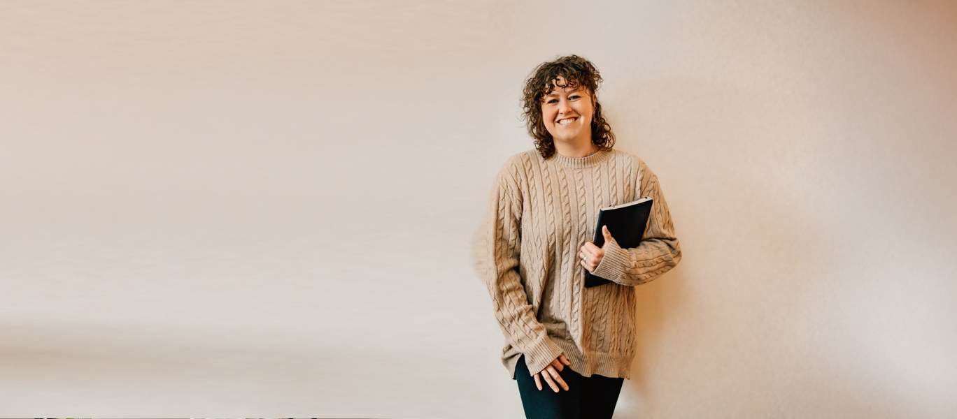 A young woman with curly hair smiling and holding a black notebook, standing against a plain beige wall.