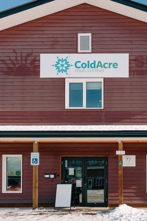 Front view of a building with a sign reading "ColdAcre Food Systems," displaying a blue logo, and a reserved wheelchair accessible parking sign outside, with snow on the ground.