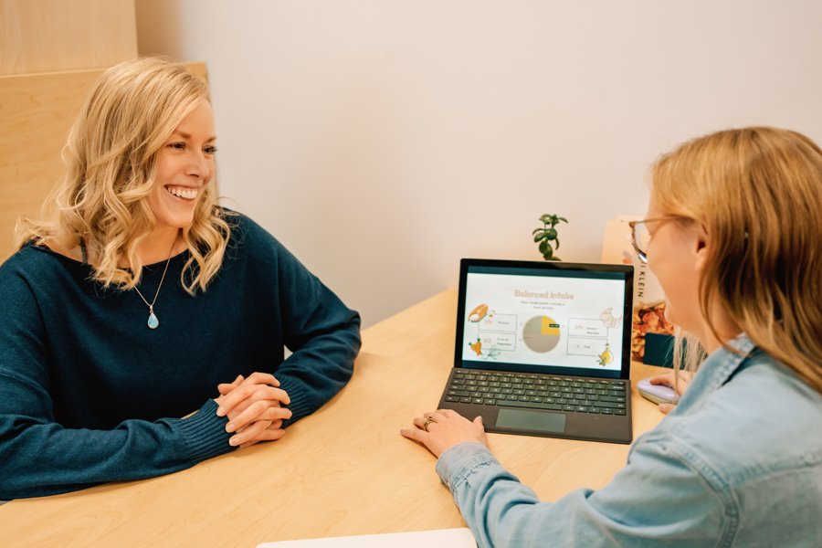 A woman with blonde hair and a woman with red hair are sitting at a wooden table in an office or meeting room. The woman on the left is smiling with her hands clasped on the table. The woman on the right, wearing glasses, is looking at a tablet that displays a presentation slide titled "Balanced Insula" with a pie chart and diagrams. There is a small plant and some books on the table.