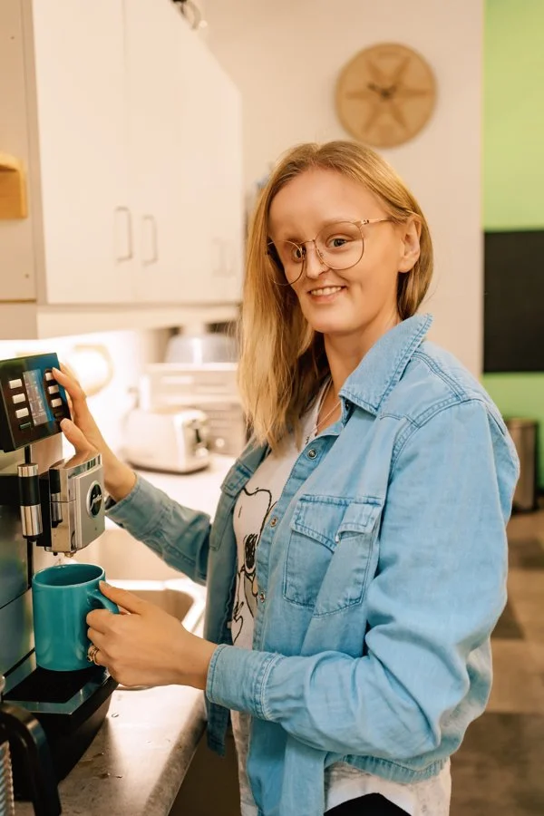 A woman with glasses and blonde hair making coffee with a coffee machine in a kitchen.