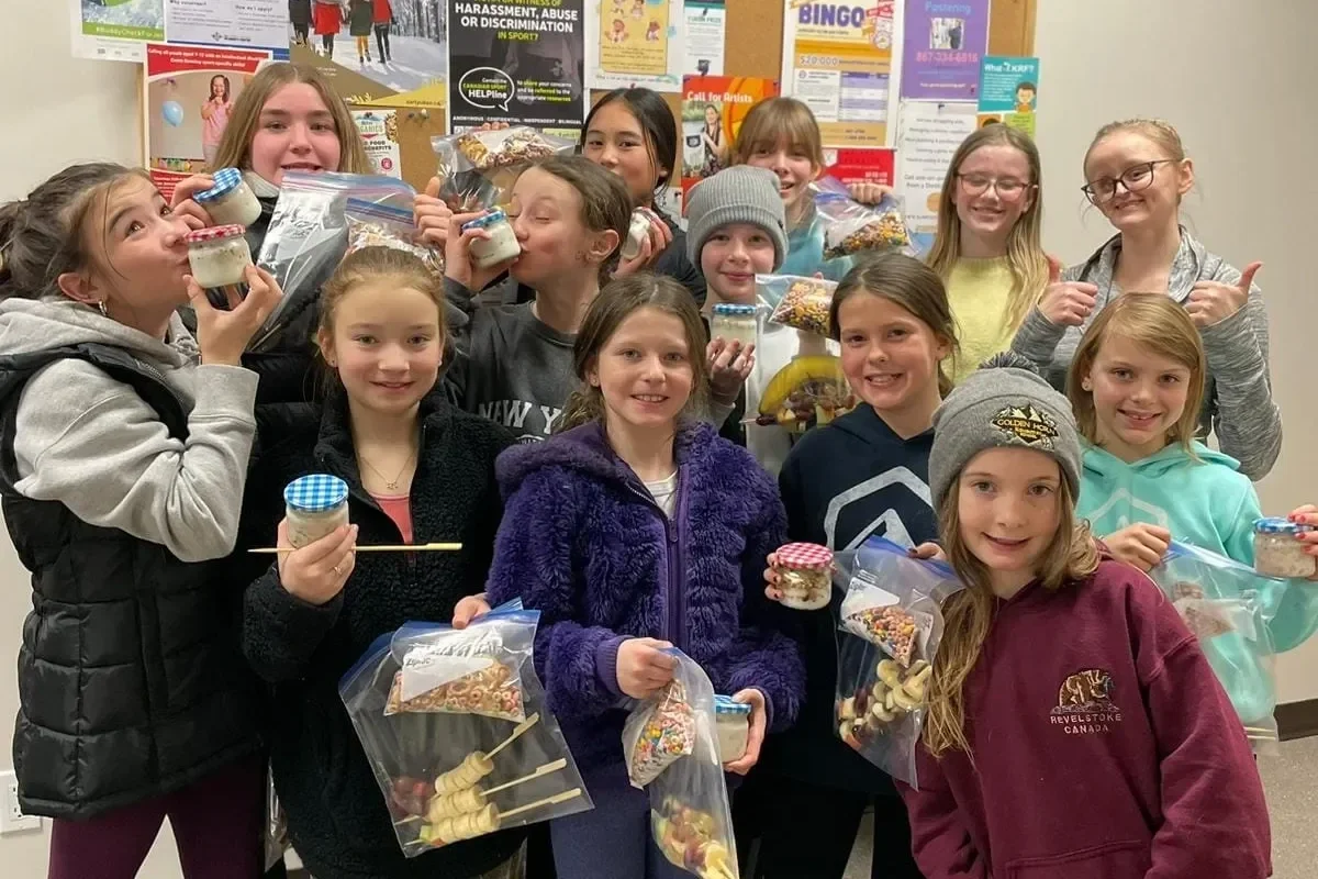 Group of 14 children smiling and holding jars of decorated cookies and bags of assorted snacks in front of a bulletin board with posters.
