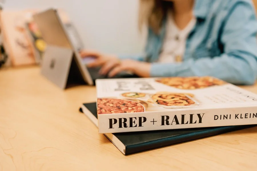 Notebook and cookbook sitting on table, with laptop/ women's hands typing in the background.