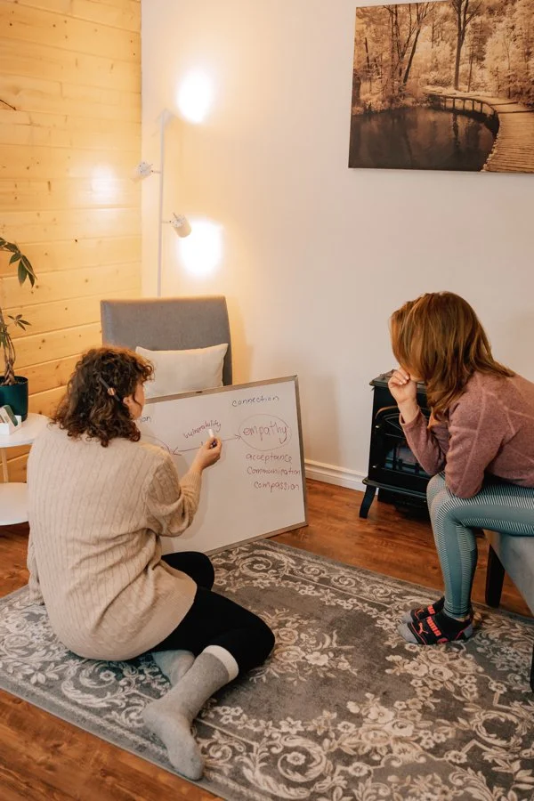 Two women engaging in a therapy or teaching session in a cozy room. One is sitting on the floor, writing on a whiteboard, while the other is sitting on a chair, listening attentively. The whiteboard has words related to empathy and connection. The room features a patterned rug, wooden wall paneling, a framed nature photo, and soft lighting.