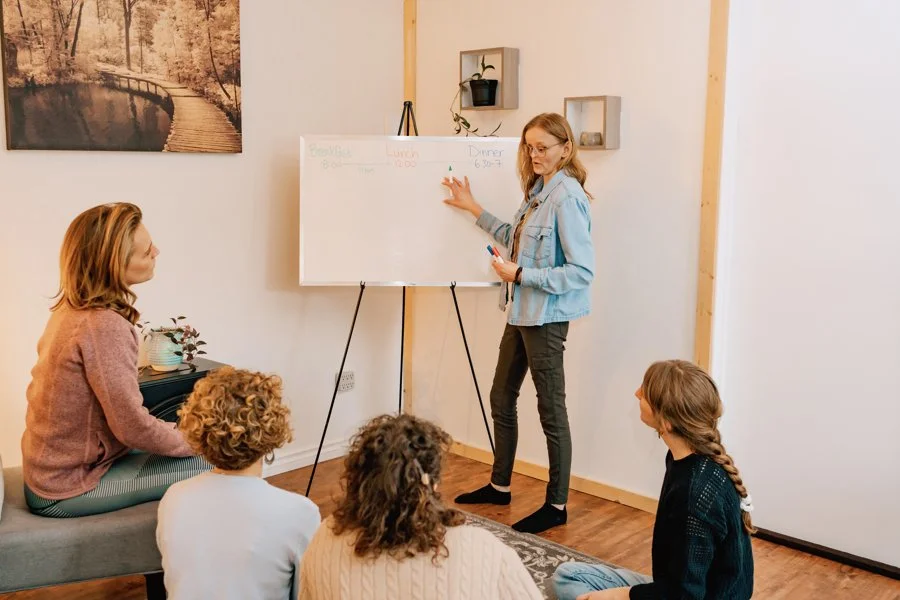 A woman gives a presentation in a room with five children seated on the floor and a sofa, near a whiteboard on a stand, with framed artwork and shelves on the wall.