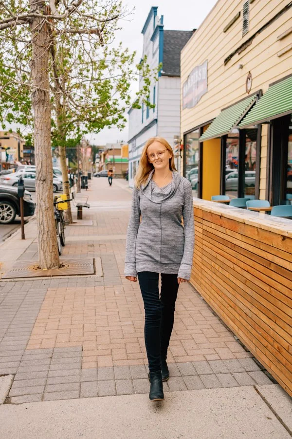 A woman with blonde hair, glasses, and a gray sweater walking on a city sidewalk.