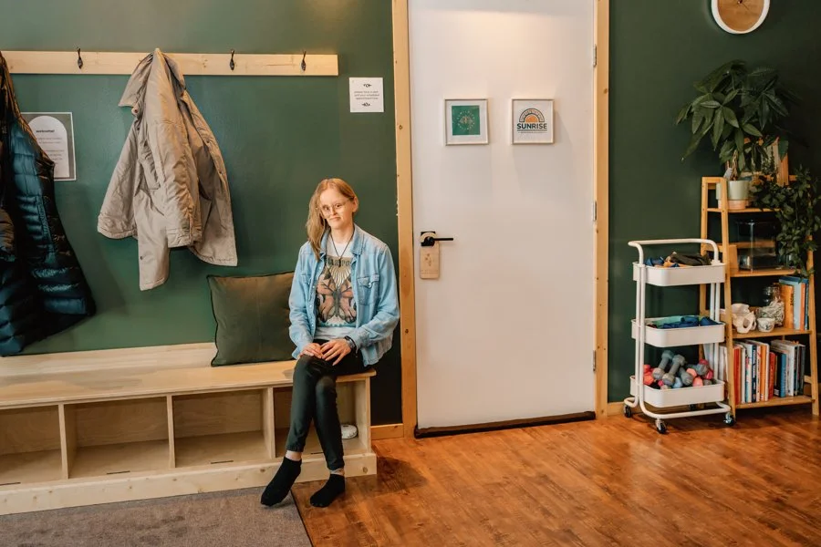 A young girl sitting on a wooden bench in a room with green and white walls, with jackets hanging on hooks behind her and a plant on a shelf to her right.