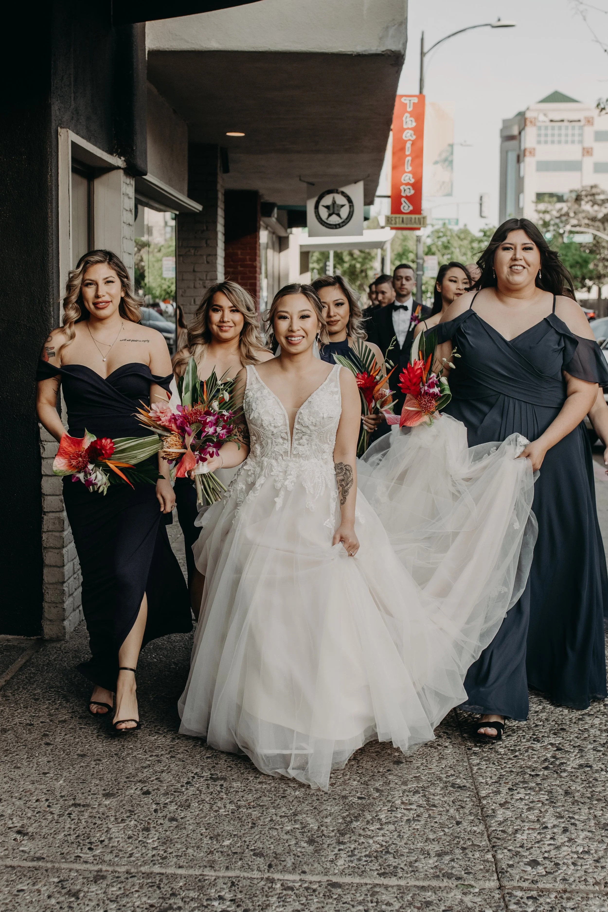 A bride in a white wedding dress accompanied by five bridesmaids in dark dresses walking together on a city sidewalk with buildings and signs in the background.
