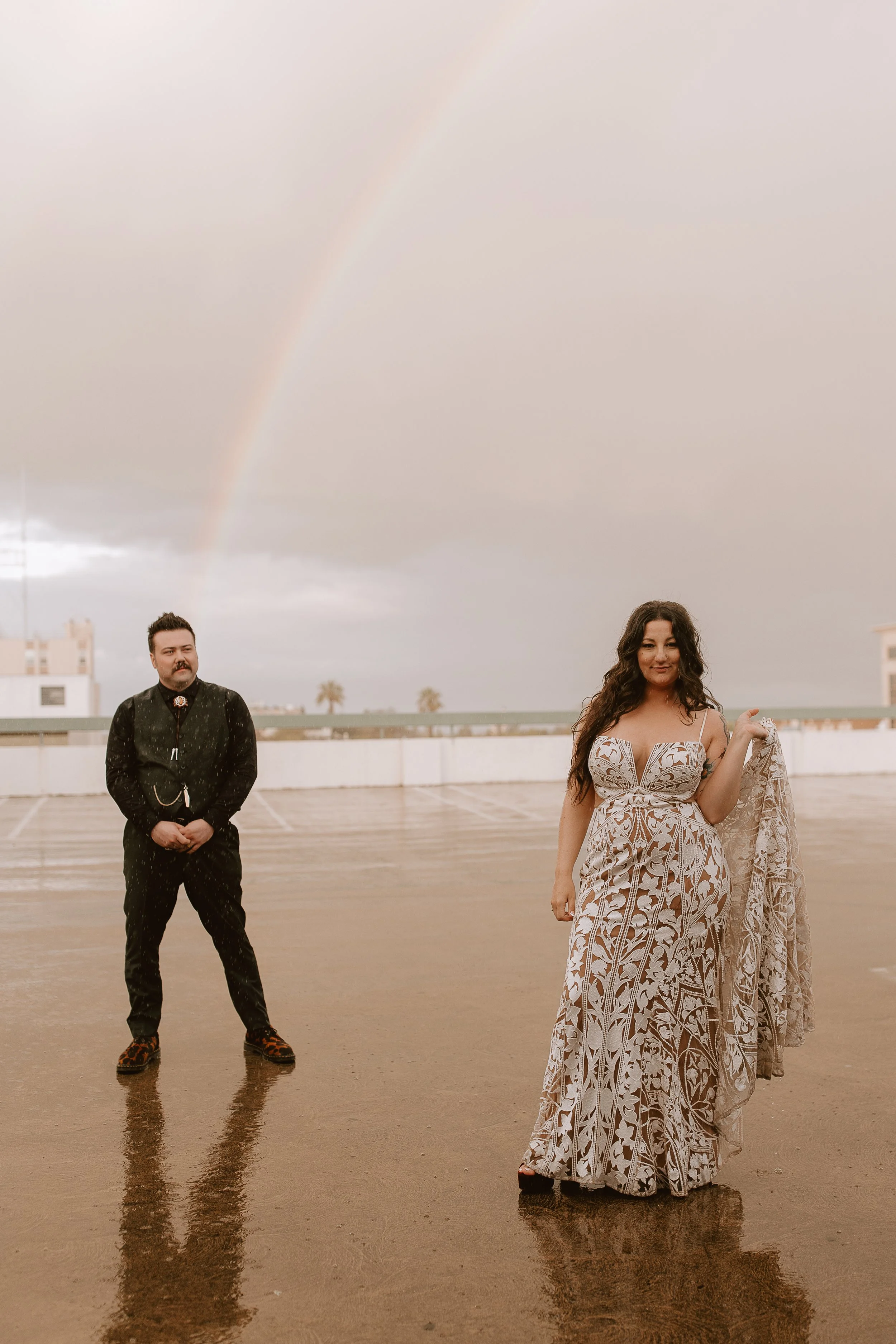 A woman in a patterned dress holding a shawl stands on a wet parking lot with a rainbow in the cloudy sky behind her. A man in black stands at a distance, under a cloudy sky with rain visible.