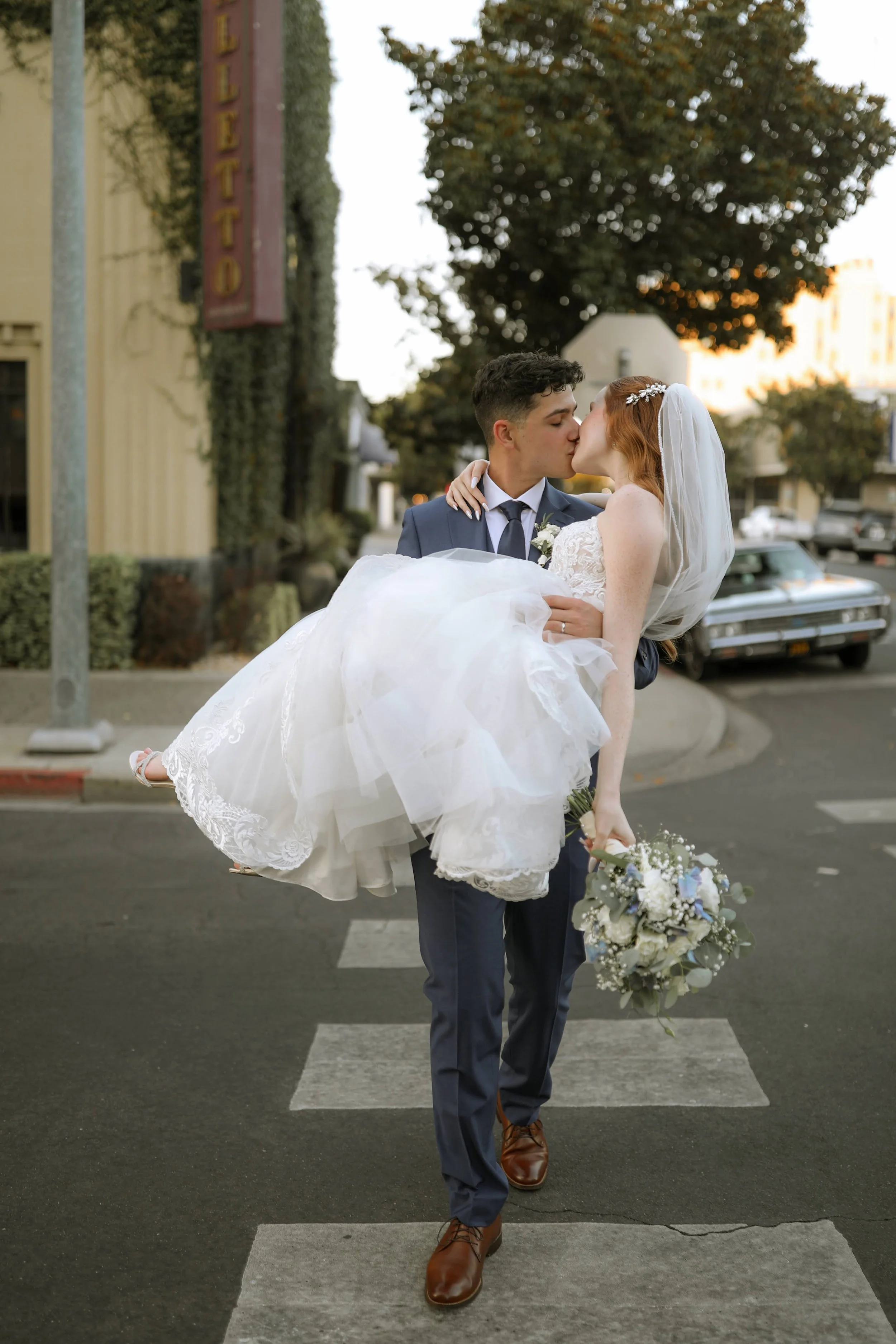 Groom in a navy suit carrying bride in a white wedding dress and veil across the street as they kiss, with vintage cars and a building with a restaurant sign in the background.