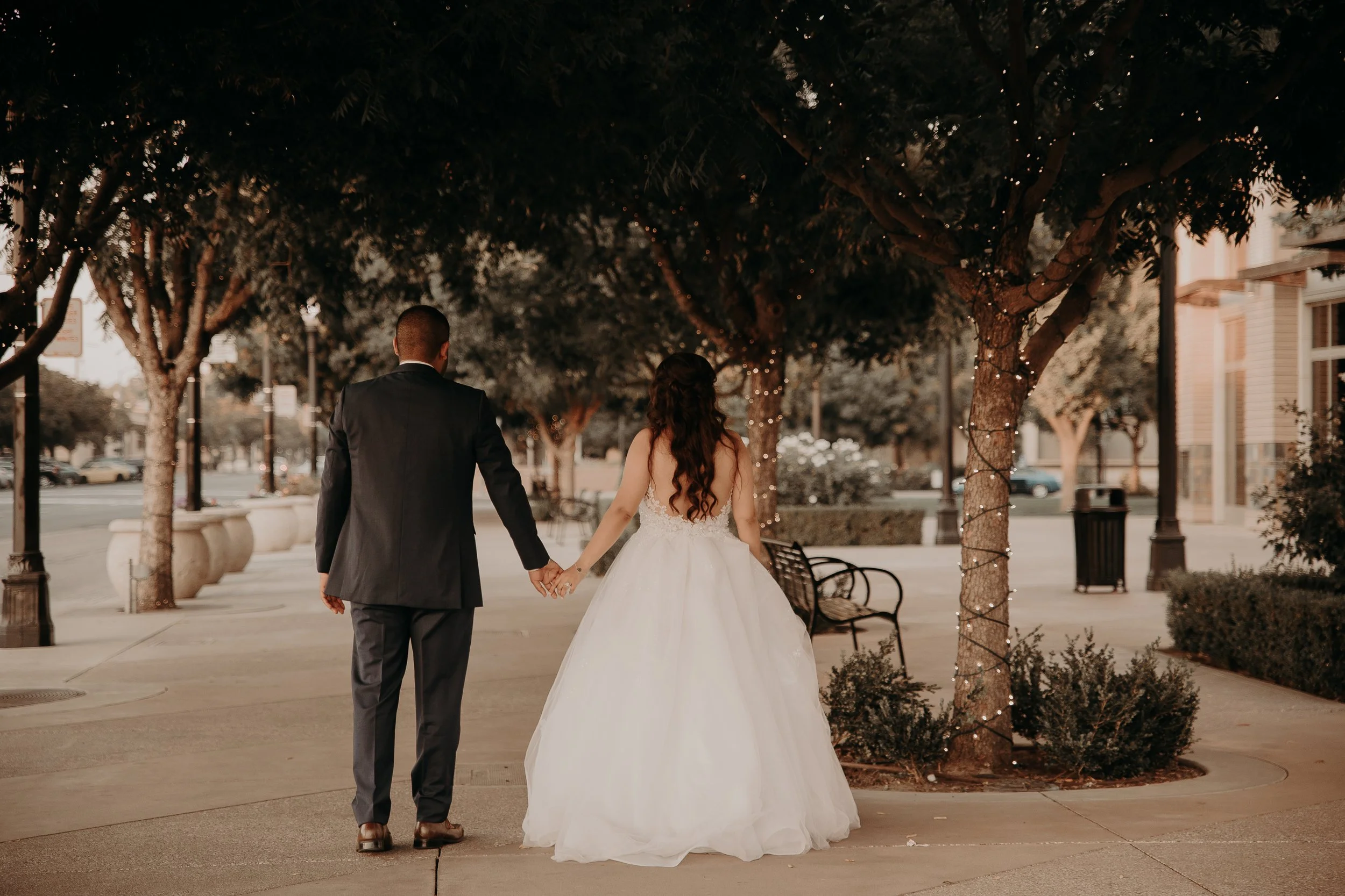 A couple dressed in wedding attire holding hands and walking down a sidewalk lined with trees, some decorated with string lights, in an outdoor urban setting.