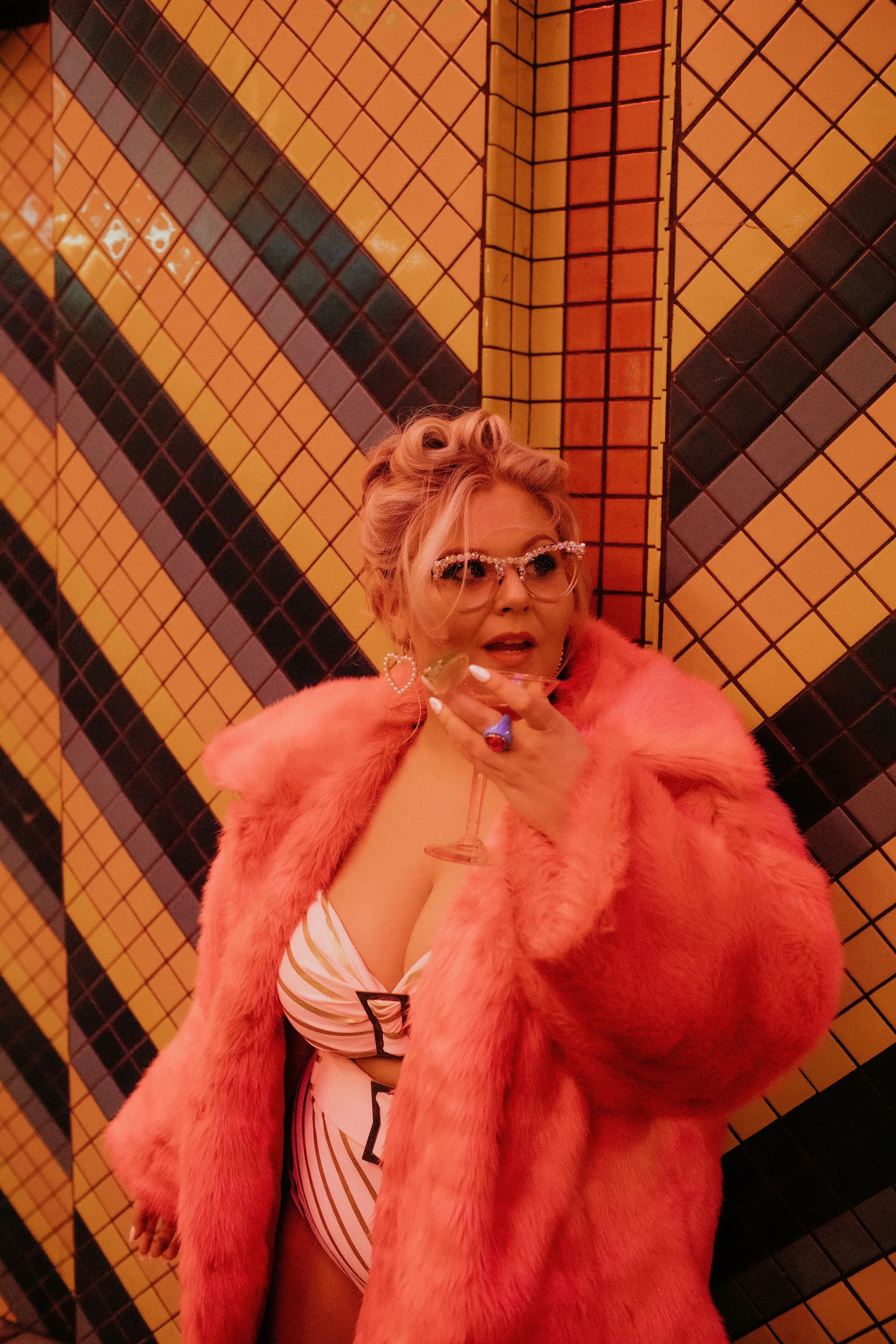 A woman wearing a pink fur coat and sunglasses, holding a glass of wine, standing in front of a colorful tiled wall.