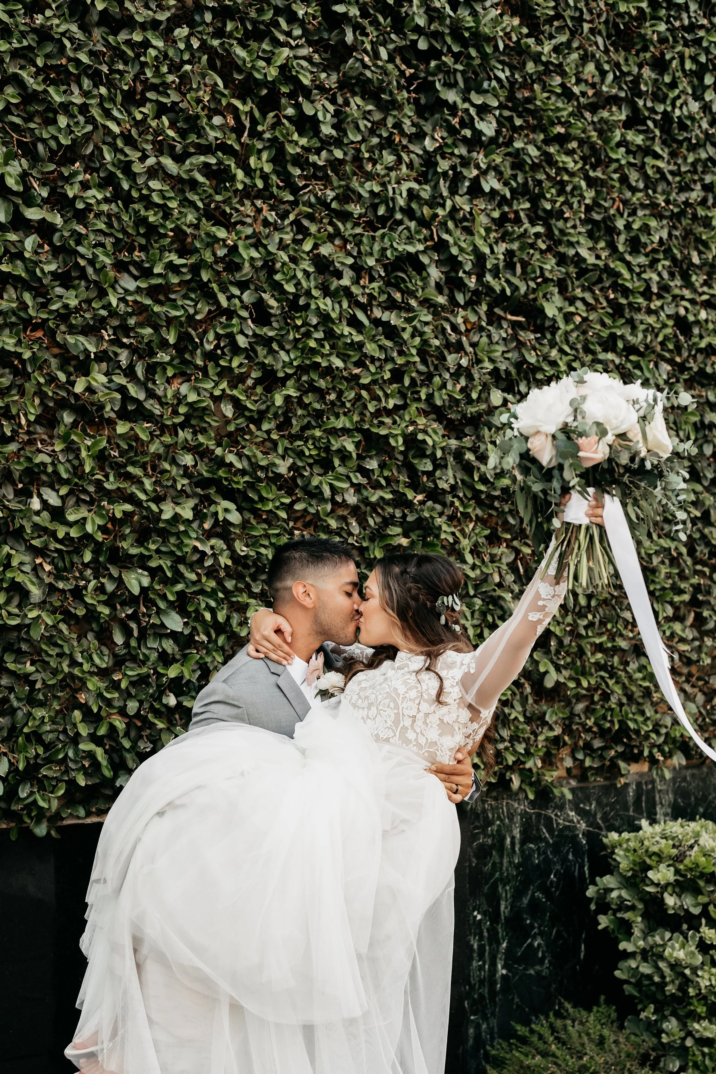 A bride and groom share a kiss outdoors, with the bride holding a large bouquet of white and blush pink flowers raised in the air, against a dense green hedge background.