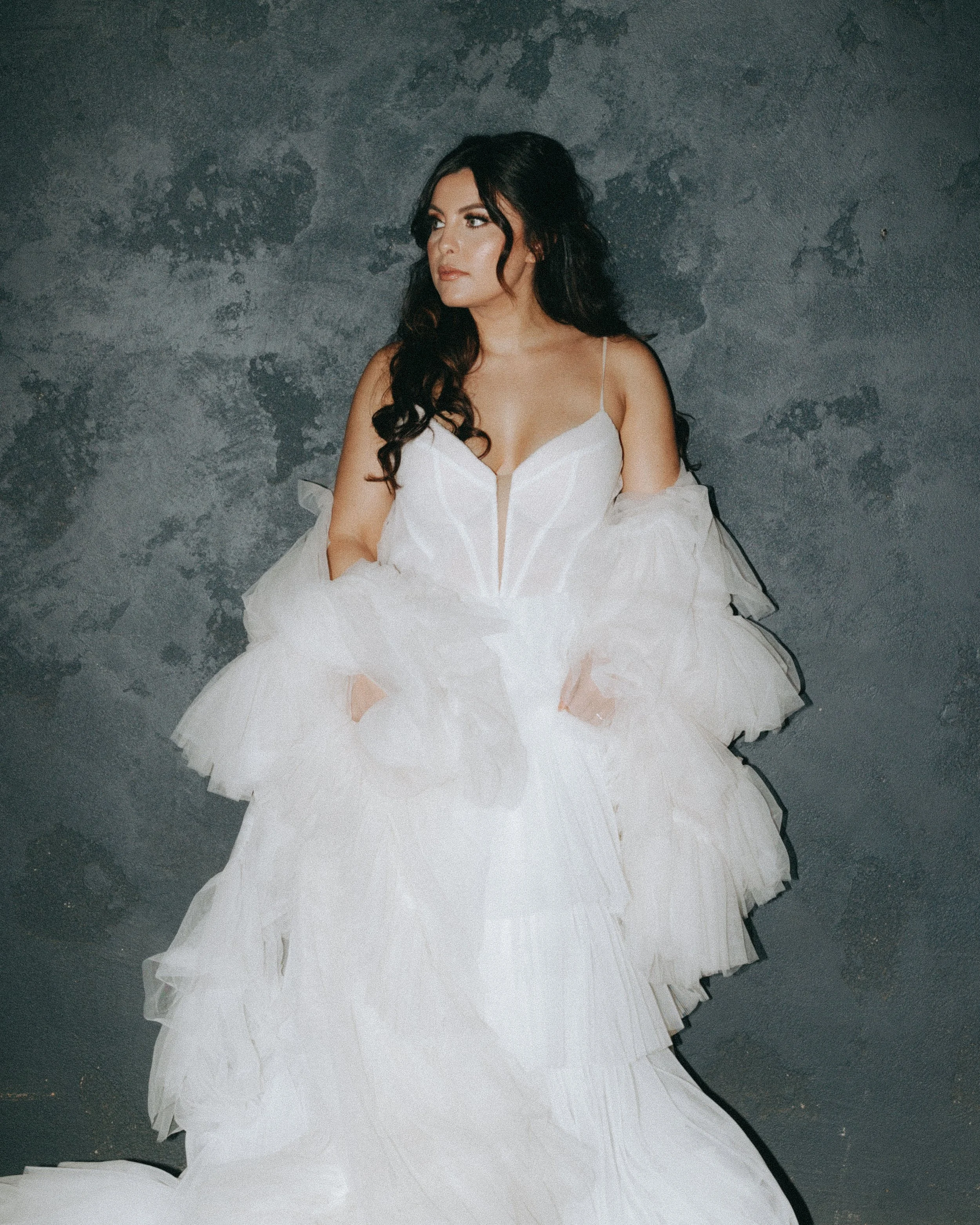 A woman with long dark hair and light skin wearing a white gown with ruffled layers and a fitted bodice, standing against a textured gray wall.