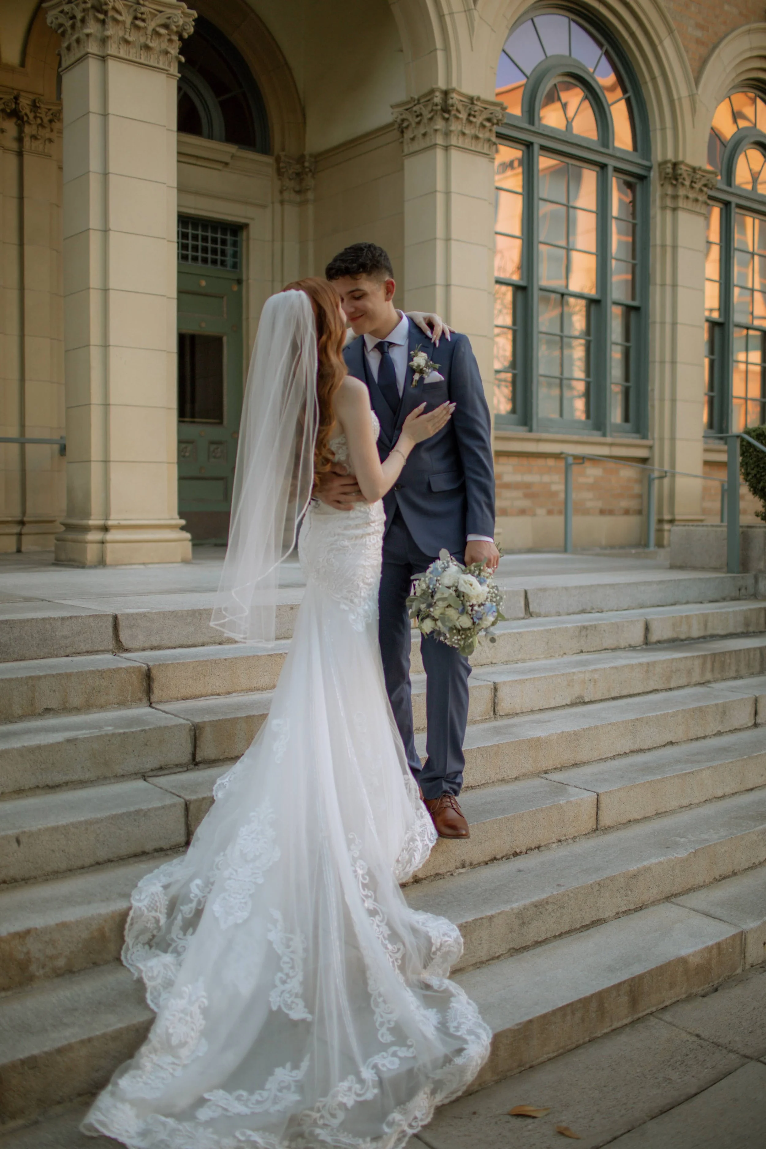 A bride and groom sharing a tender moment on the steps outside a historic building during sunset. The bride wears a white lace wedding gown with a long train and a veil, while the groom is dressed in a dark suit holding a bouquet of flowers.