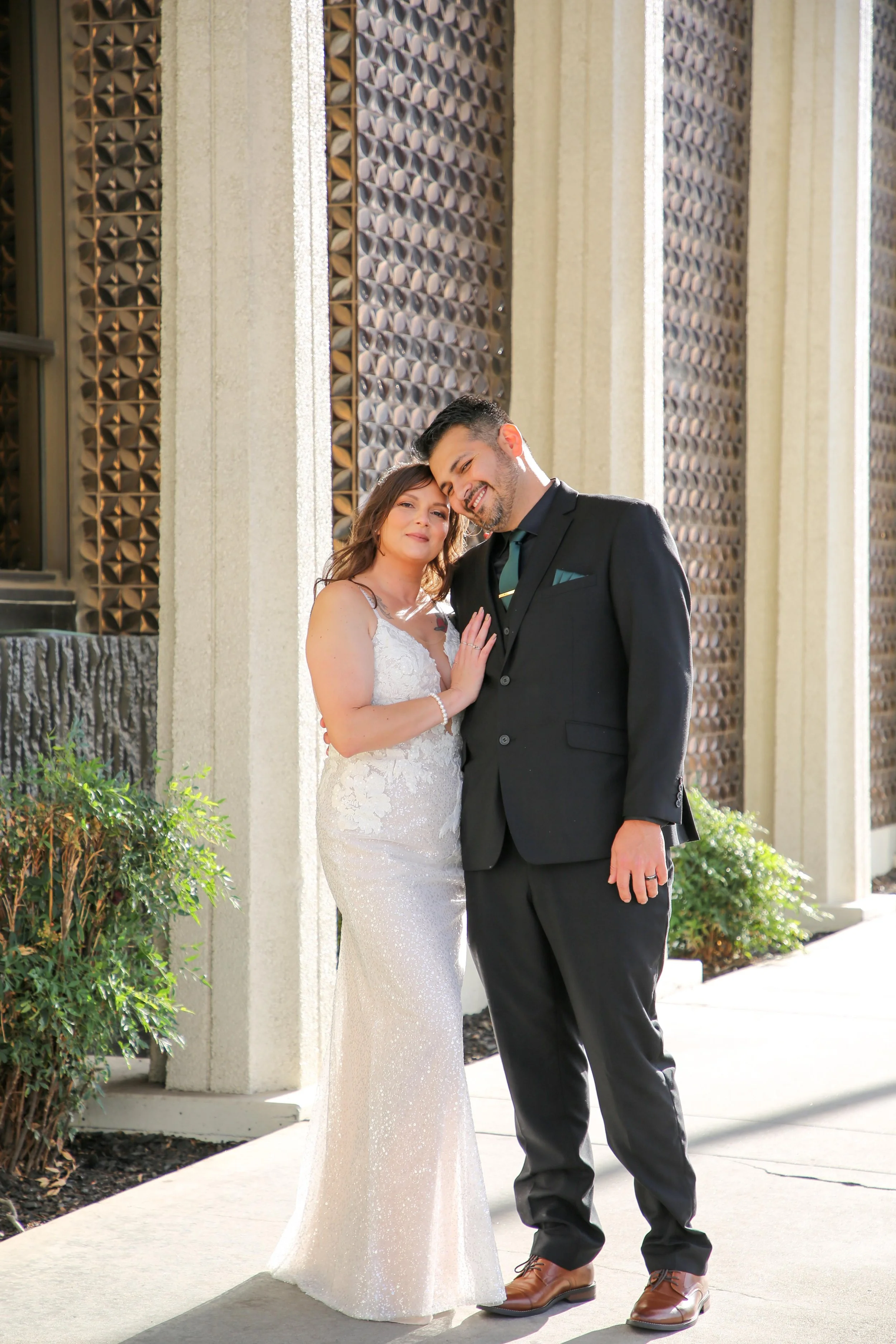 A woman in a white, sparkly lace wedding dress and a man in a black suit and teal tie, standing close together outdoors, smiling, against a geometric metal wall background with green bushes.