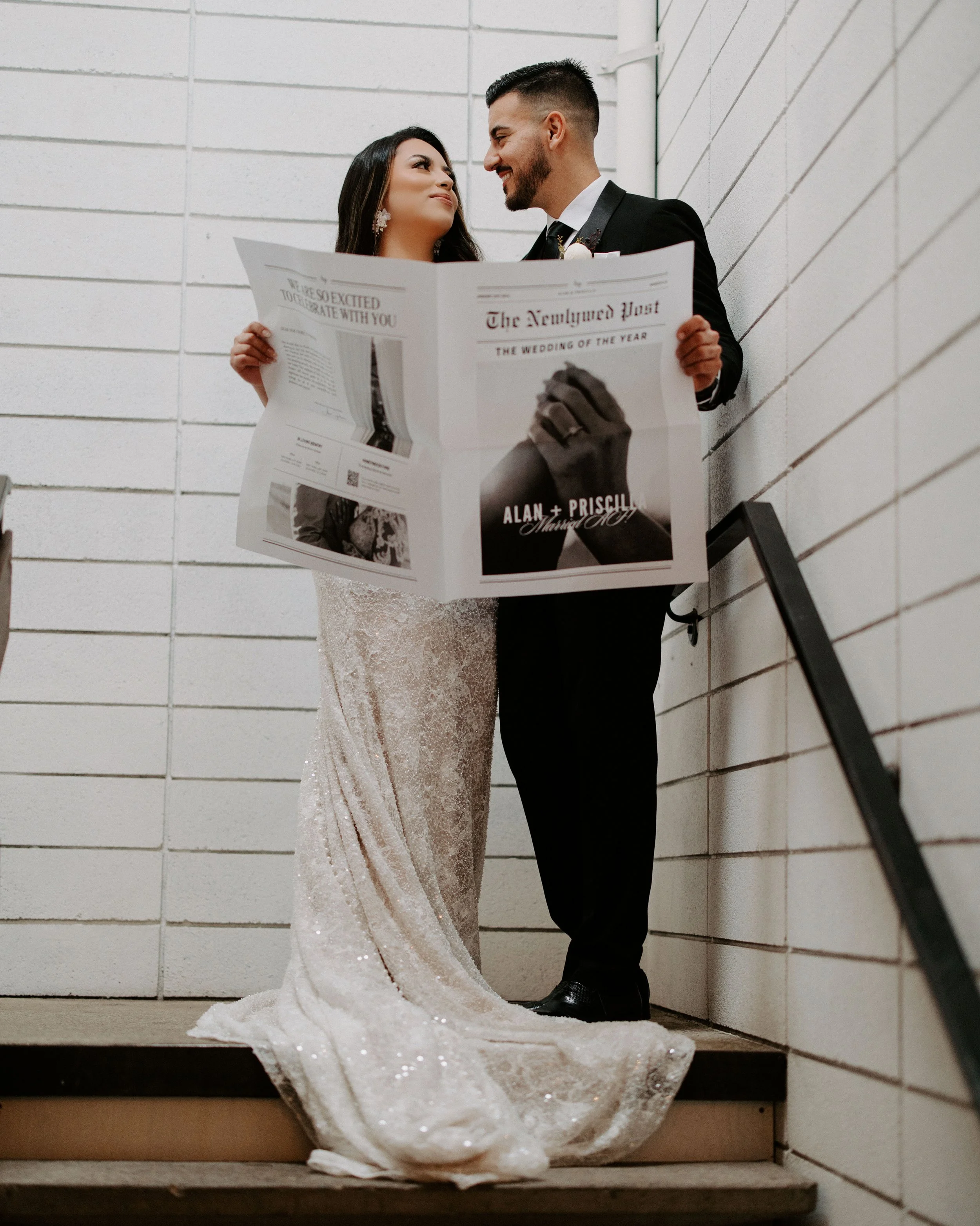 A bride and groom in wedding attire standing close together, holding a newspaper with the headline "The Newlign Post" and smiling at each other against a brick wall.