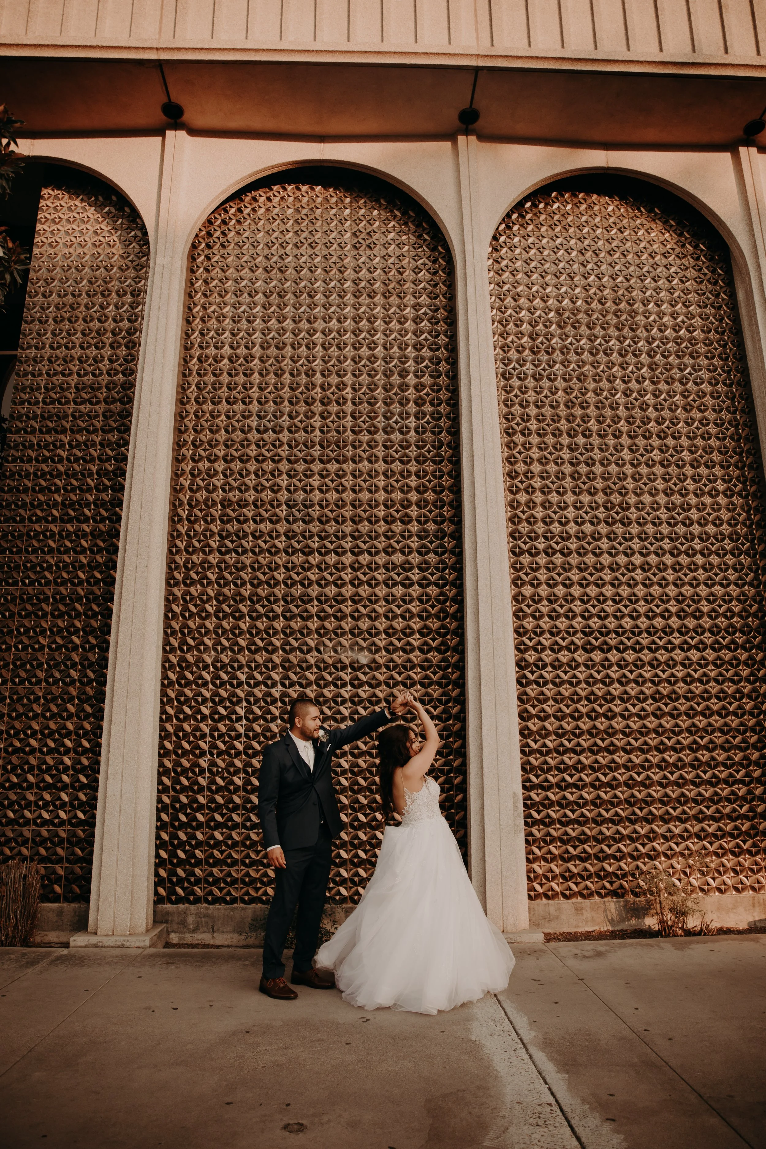 A newlywed couple dancing outside in front of a large decorative wall. The bride is wearing a white wedding gown and the groom a dark suit. They are holding hands with the groom twirling the bride.
