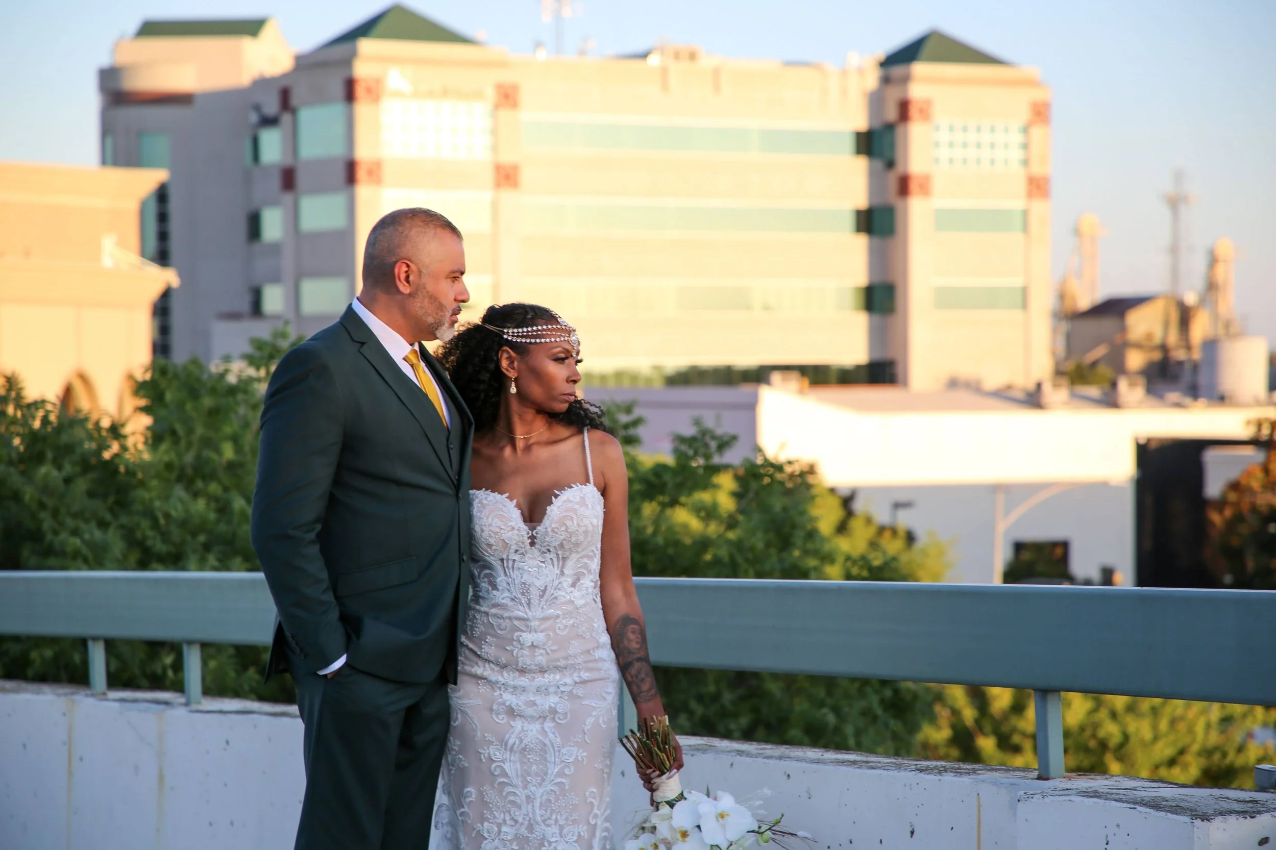 A newly married couple stands outdoors on a rooftop at sunset, with downtown buildings in the background. The bride is in a white lace wedding dress holding a bouquet, and the groom is in a black suit with a yellow tie, both looking contemplative.