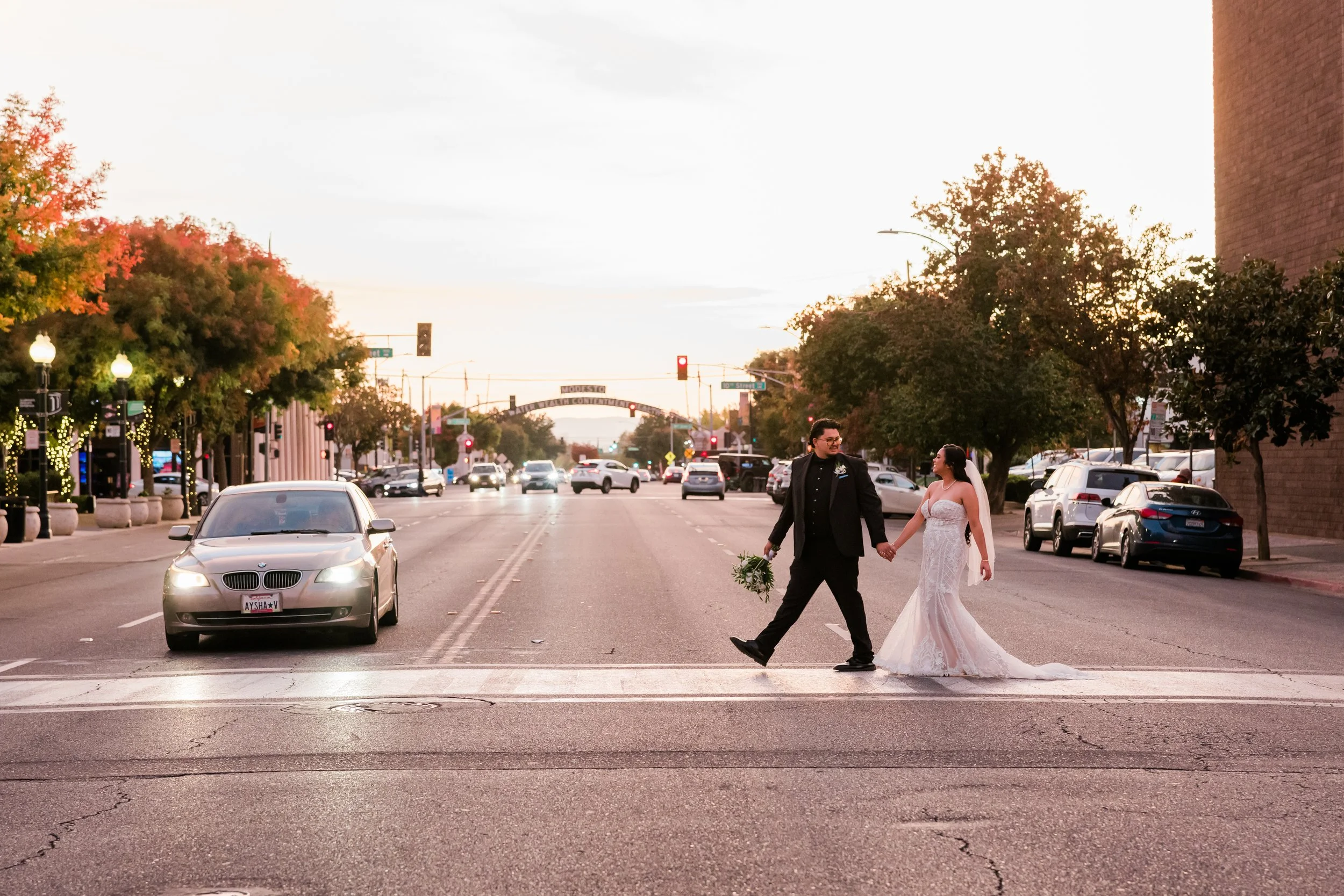 A bride and groom holding hands and crossing the street at sunset, with cars and trees in the background.