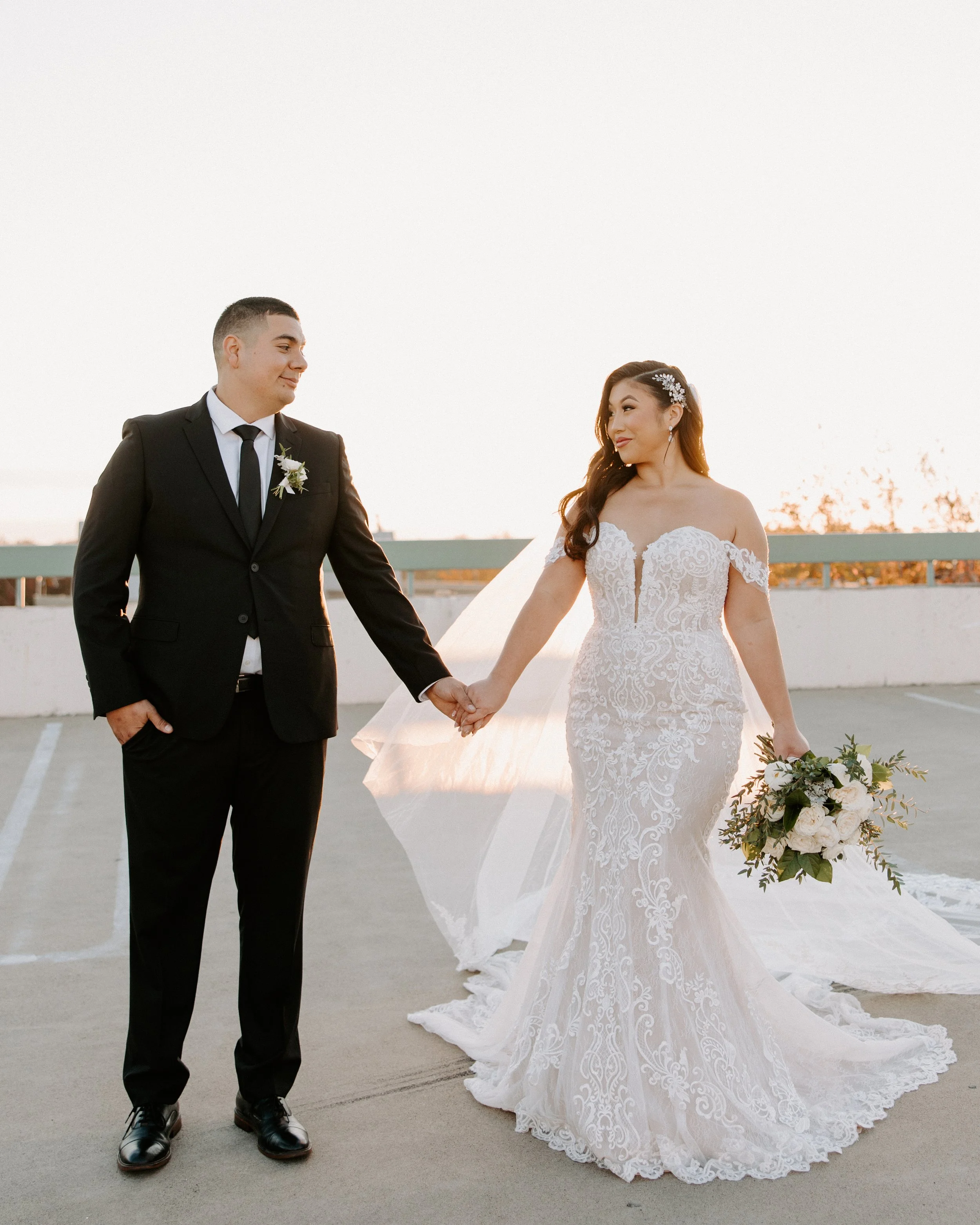 A bride and groom holding hands on a rooftop parking lot during sunset, the bride in a lace wedding dress holding a bouquet and the groom in a black suit with a white shirt and black tie.