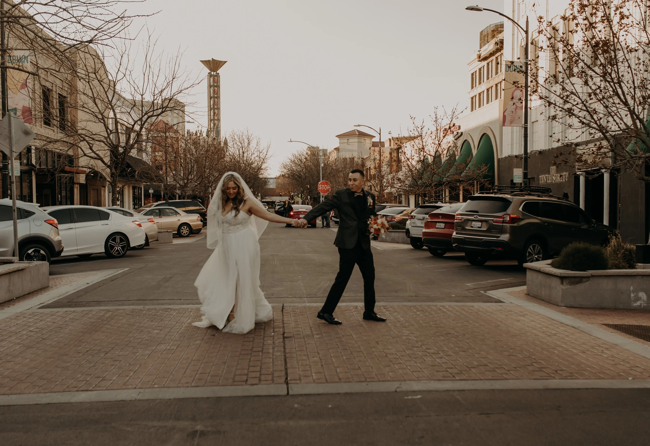 A bride and groom holding hands and dancing in an urban street during sunset, with parked cars and leafless trees in the background.