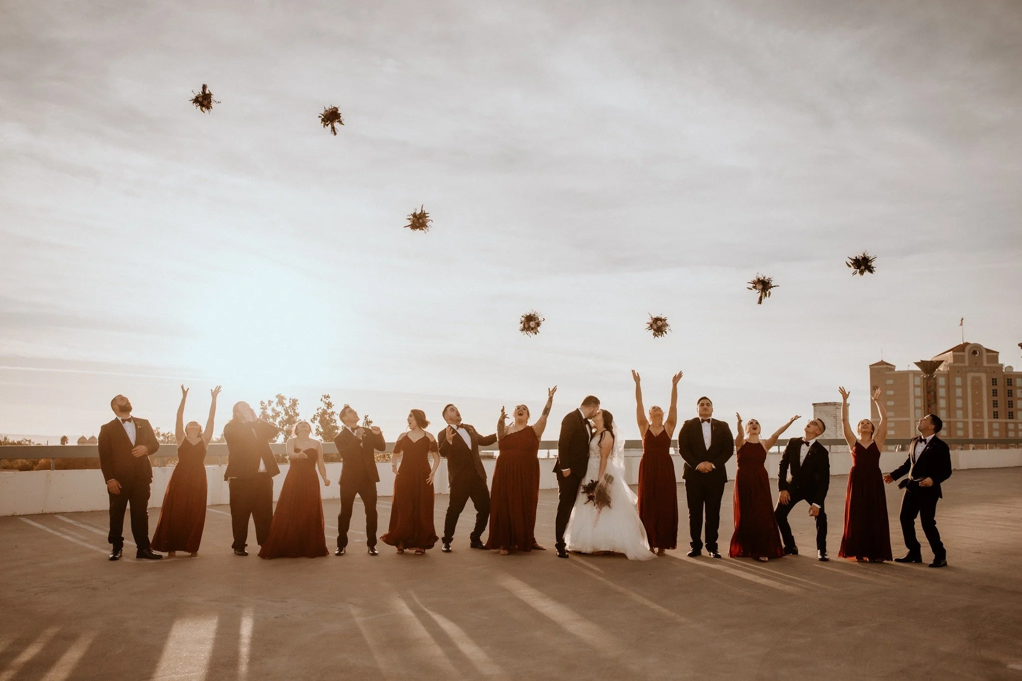 Wedding party on a rooftop releasing flower bouquets into the sky at sunset, including the bride and groom surrounded by bridesmaids and groomsmen in formal attire.