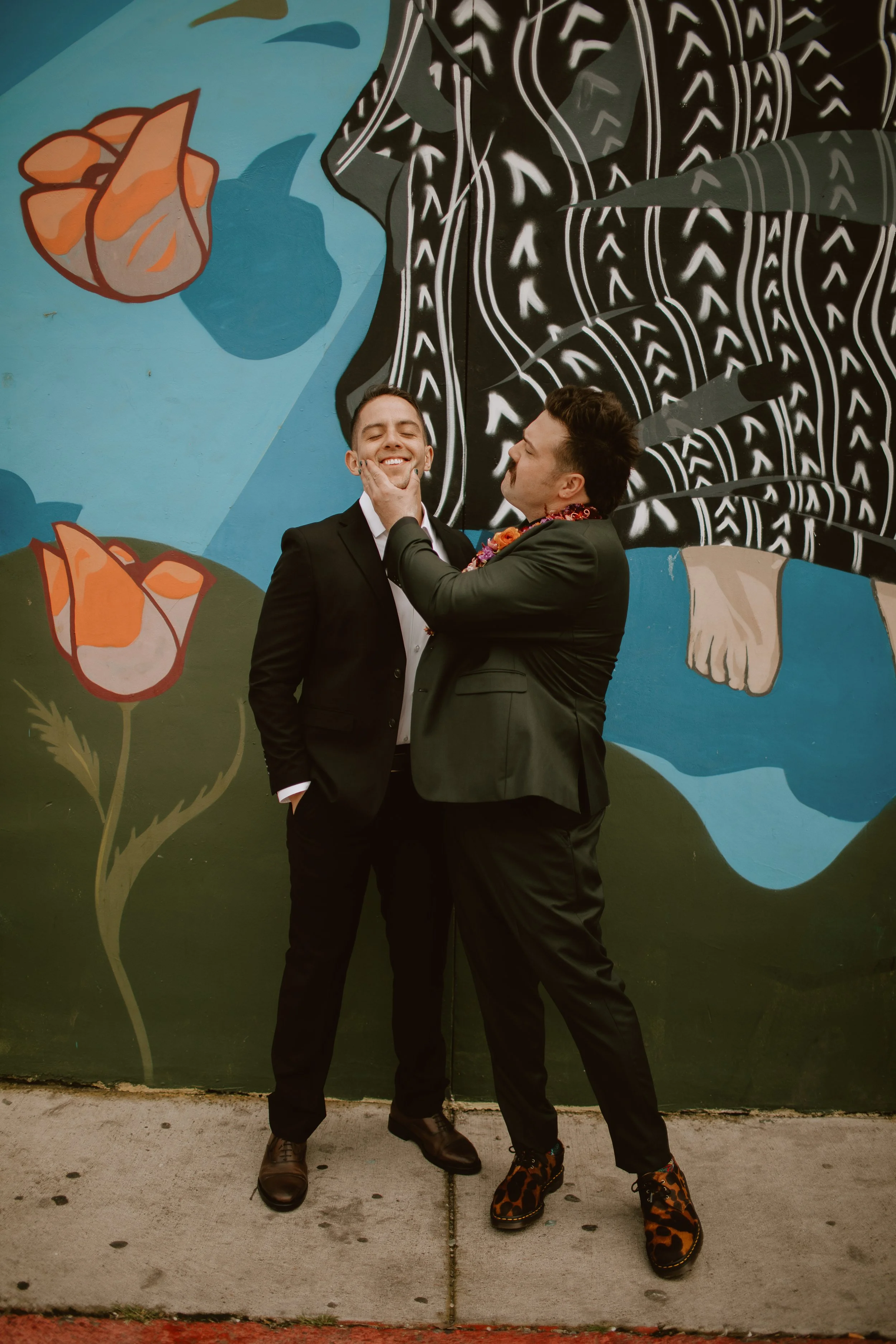 Two men in suits, one smiling and the other playfully holding his face, standing in front of a colorful mural with flowers and abstract patterns.