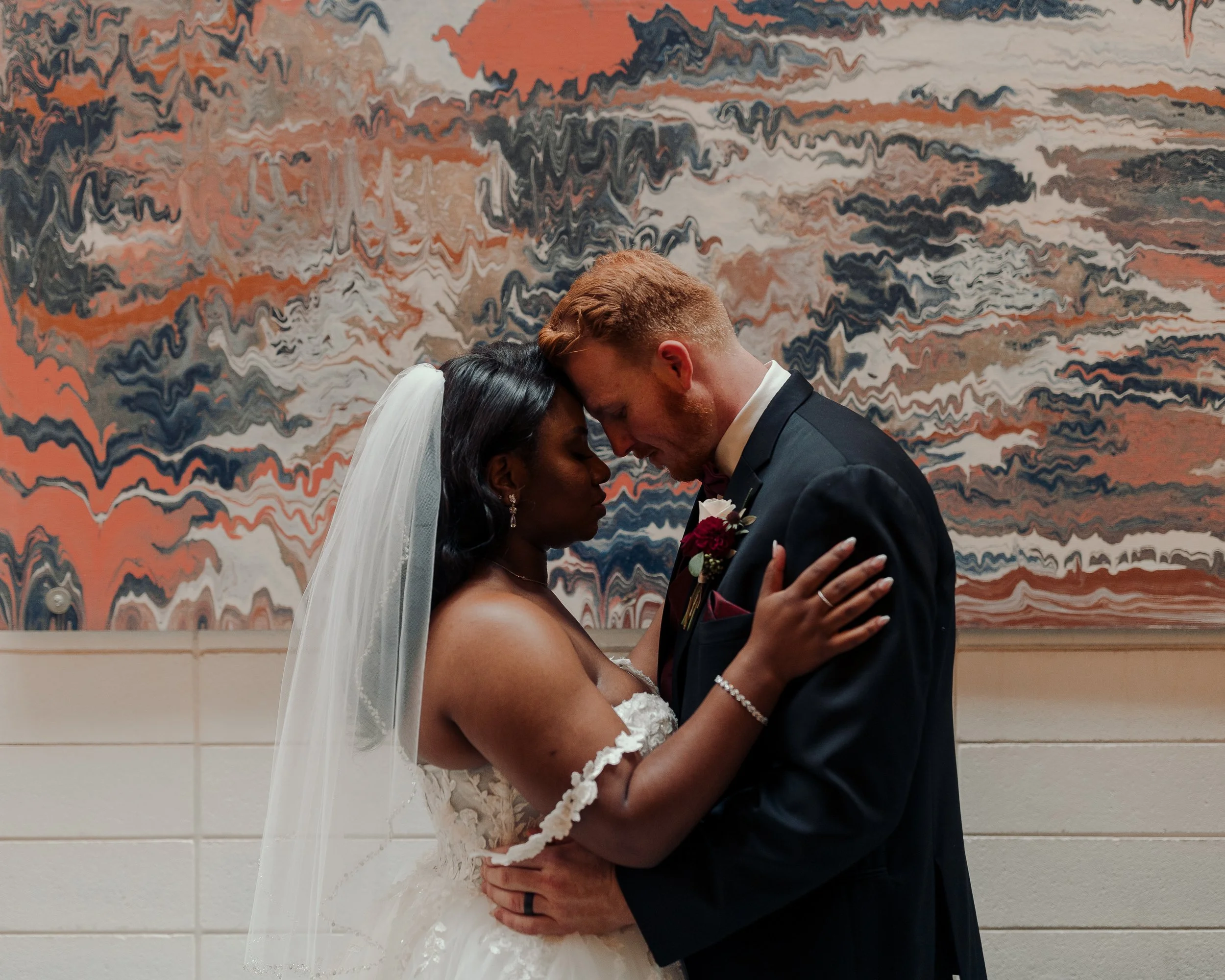 A bride and groom standing close, touching foreheads with eyes closed, in front of an abstract patterned wall, during their wedding.