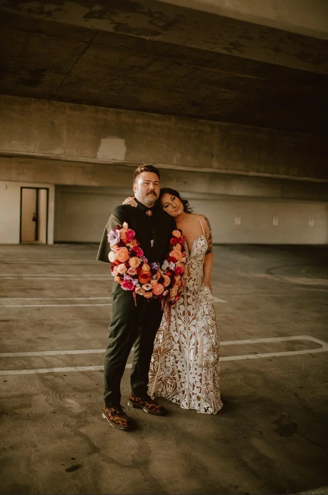 A man and woman standing close together in a parking garage, with the man wearing a black shirt and pants, decorated with a floral lei, and the woman in a white, intricate patterned dress. The scene appears warm and intimate.