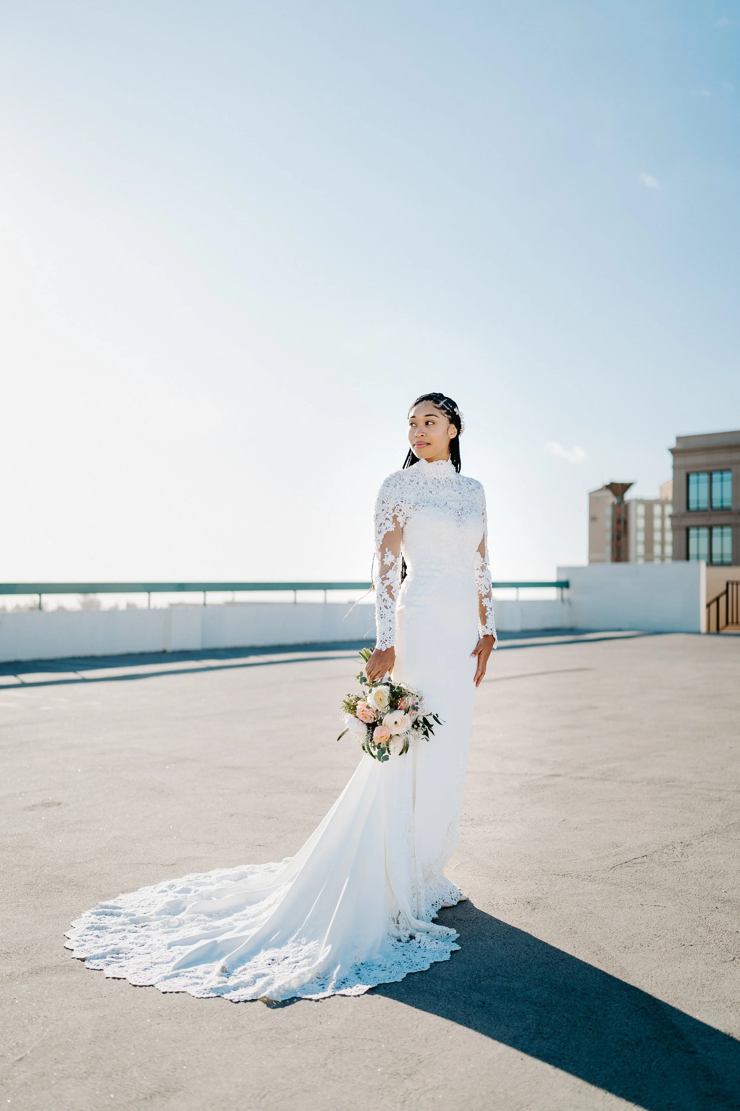 Bride in a white wedding dress holding a bouquet of flowers on a rooftop with a city skyline in the background.