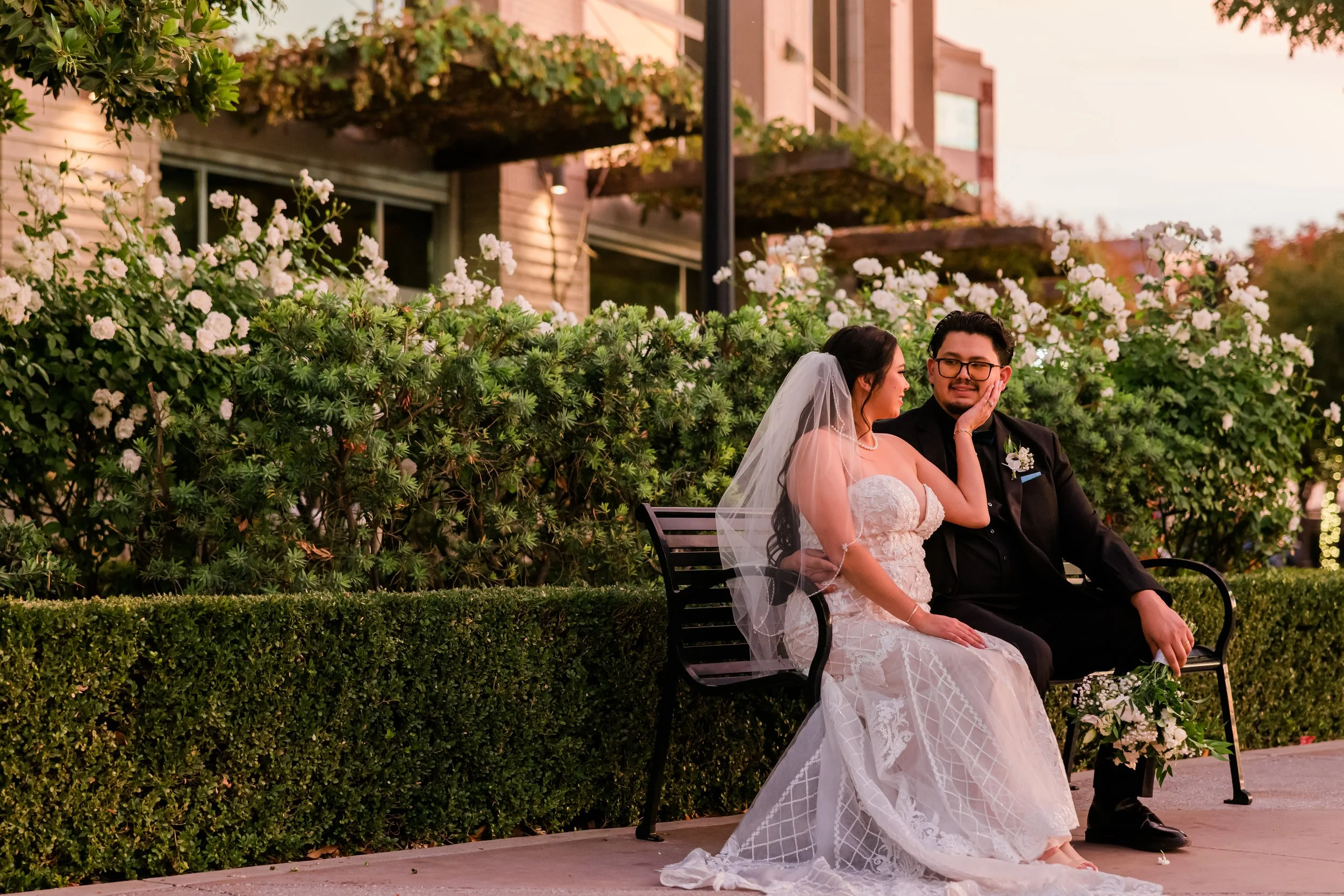 A bride and groom sitting on a bench, smiling and touching faces, outside during sunset, with lush greenery and white flowers in the background.