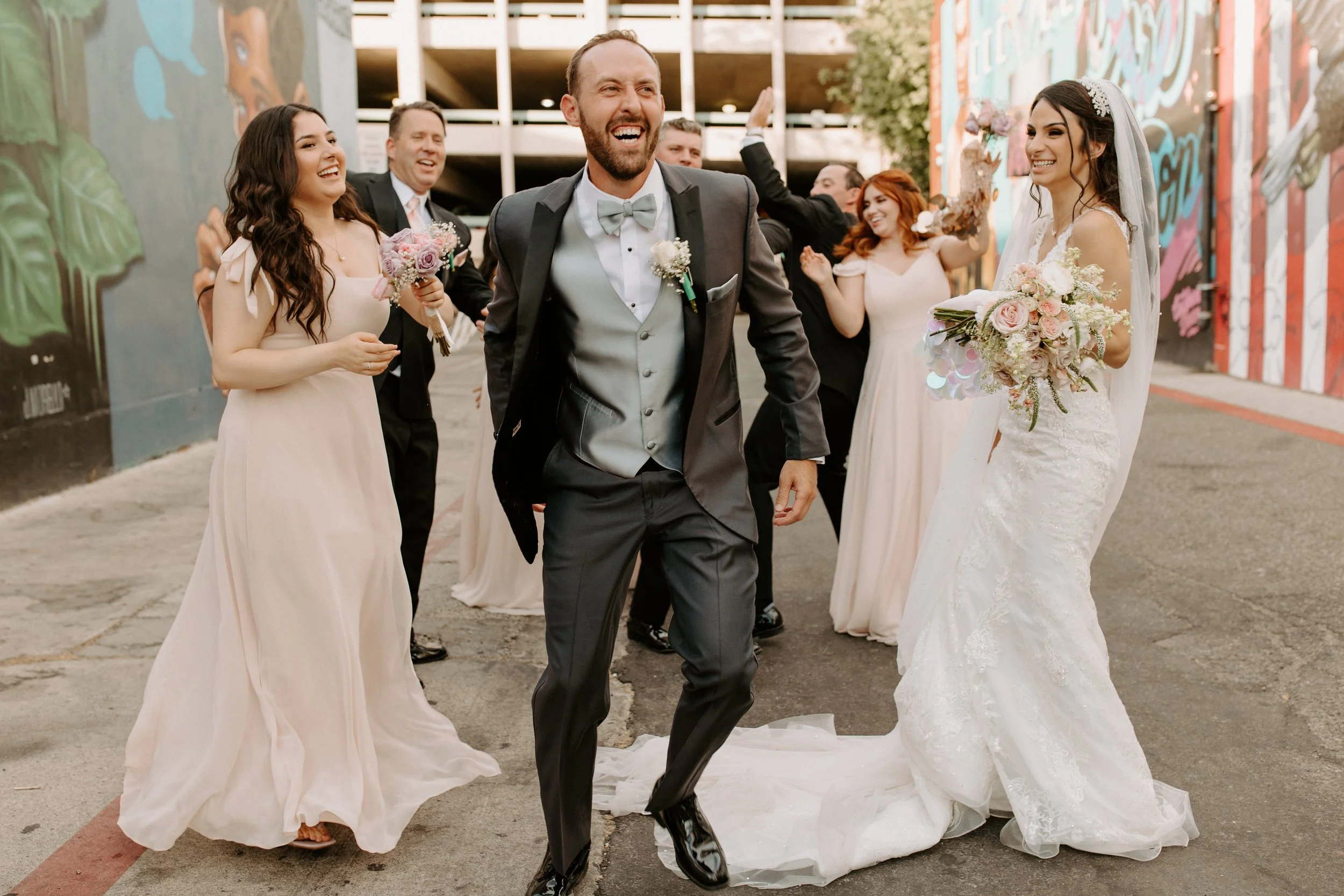 A group of wedding guests celebrating outdoors, with a bride holding a bouquet, a groom in a tuxedo, and bridesmaids in light pink dresses, all smiling and laughing.