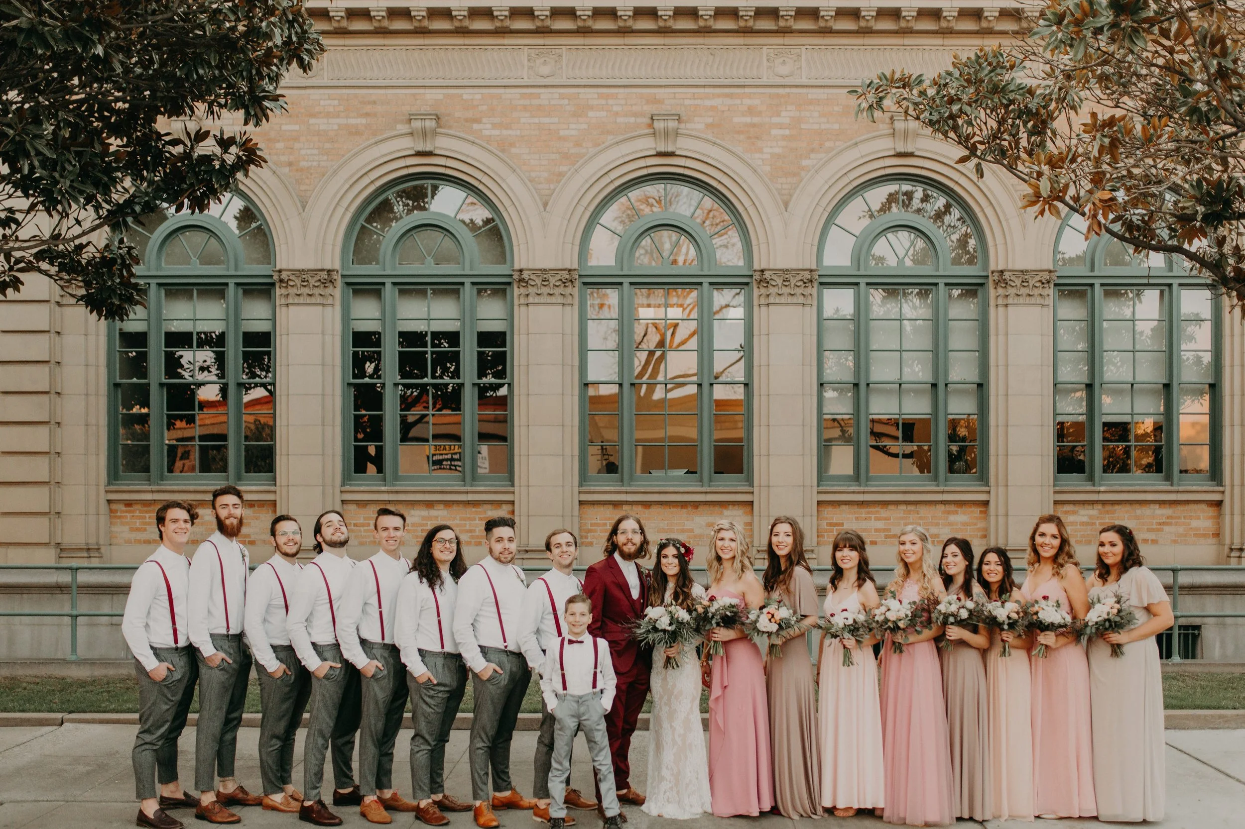 A wedding party of fourteen people, including the bride and groom, standing in front of a historic brick building with large arched windows, during sunset.