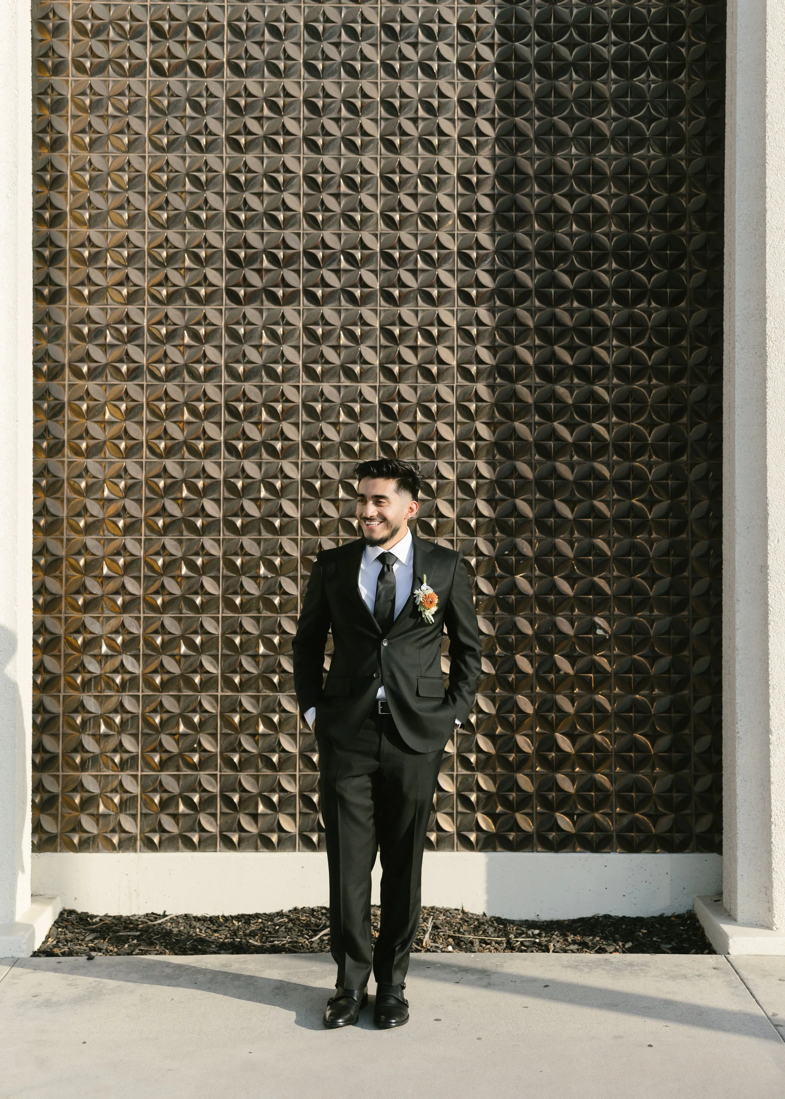 A man in a black suit with a boutonniere, white shirt, and black tie standing outside in front of a decorative metal wall, smiling with hands in pockets.