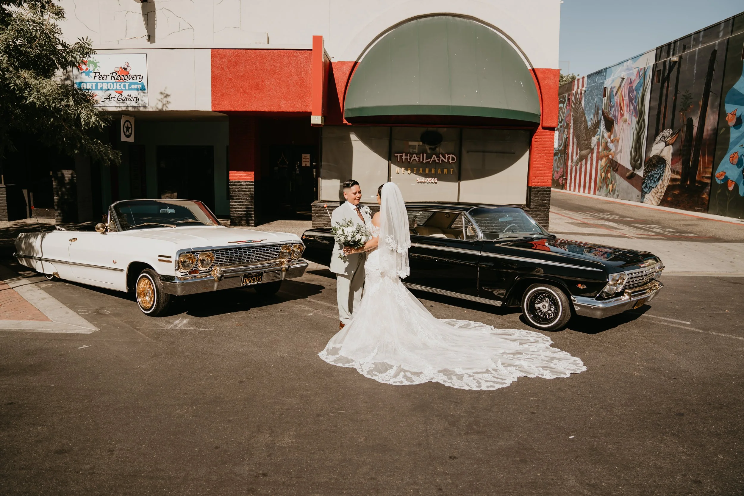 A bride and groom in wedding attire standing between two vintage cars outside a building with a sign that reads "Thailand Restaurant." The bride is wearing a long, white lace wedding gown and veil, holding a bouquet, and the groom is dressed in a lig