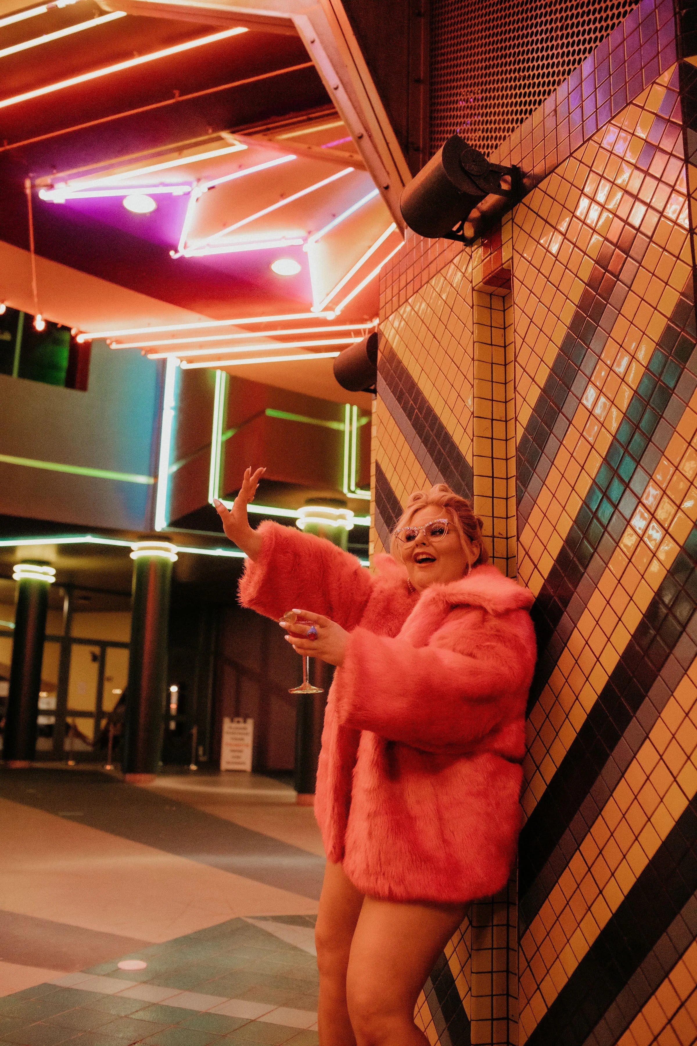 A woman in a pink fur coat and glasses smiling and holding a wine glass, standing against a brightly lit, colorful neon-themed background.