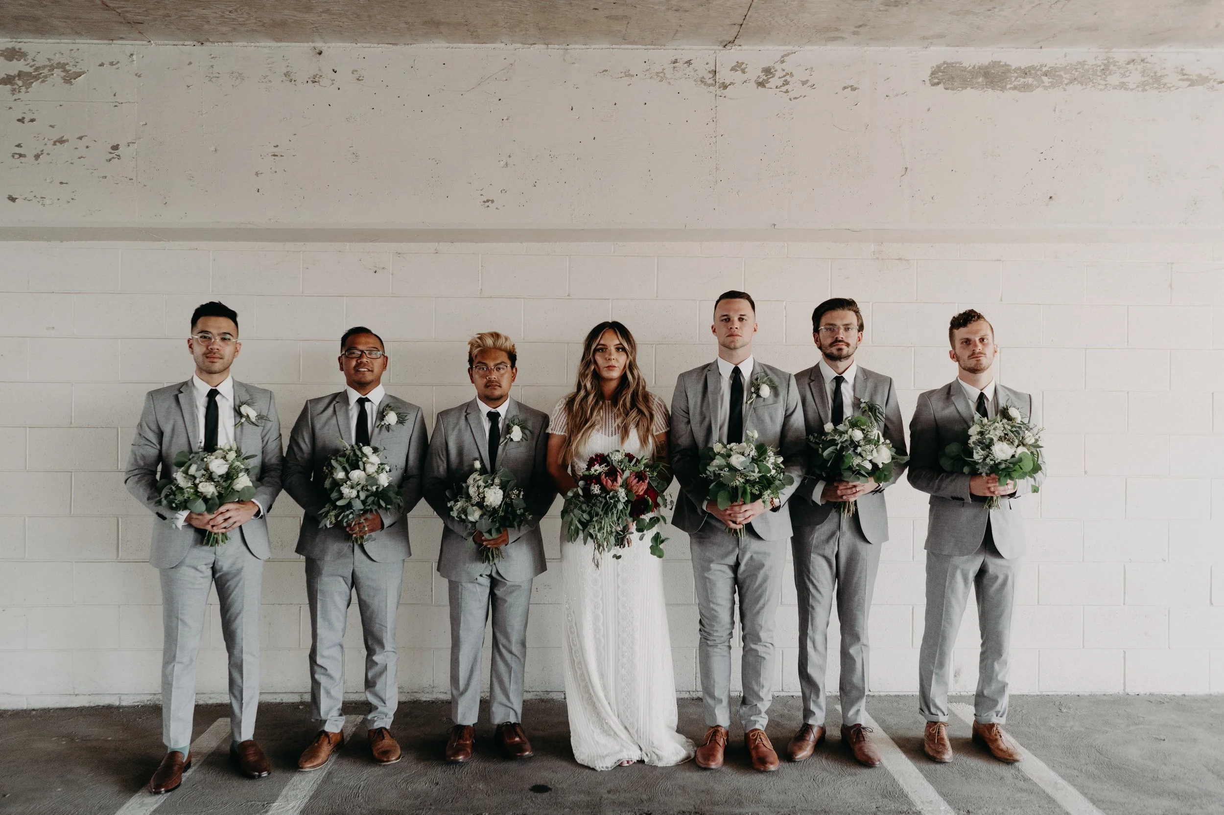 Bride and six groomsmen standing in a line holding floral bouquets in front of a white brick wall.