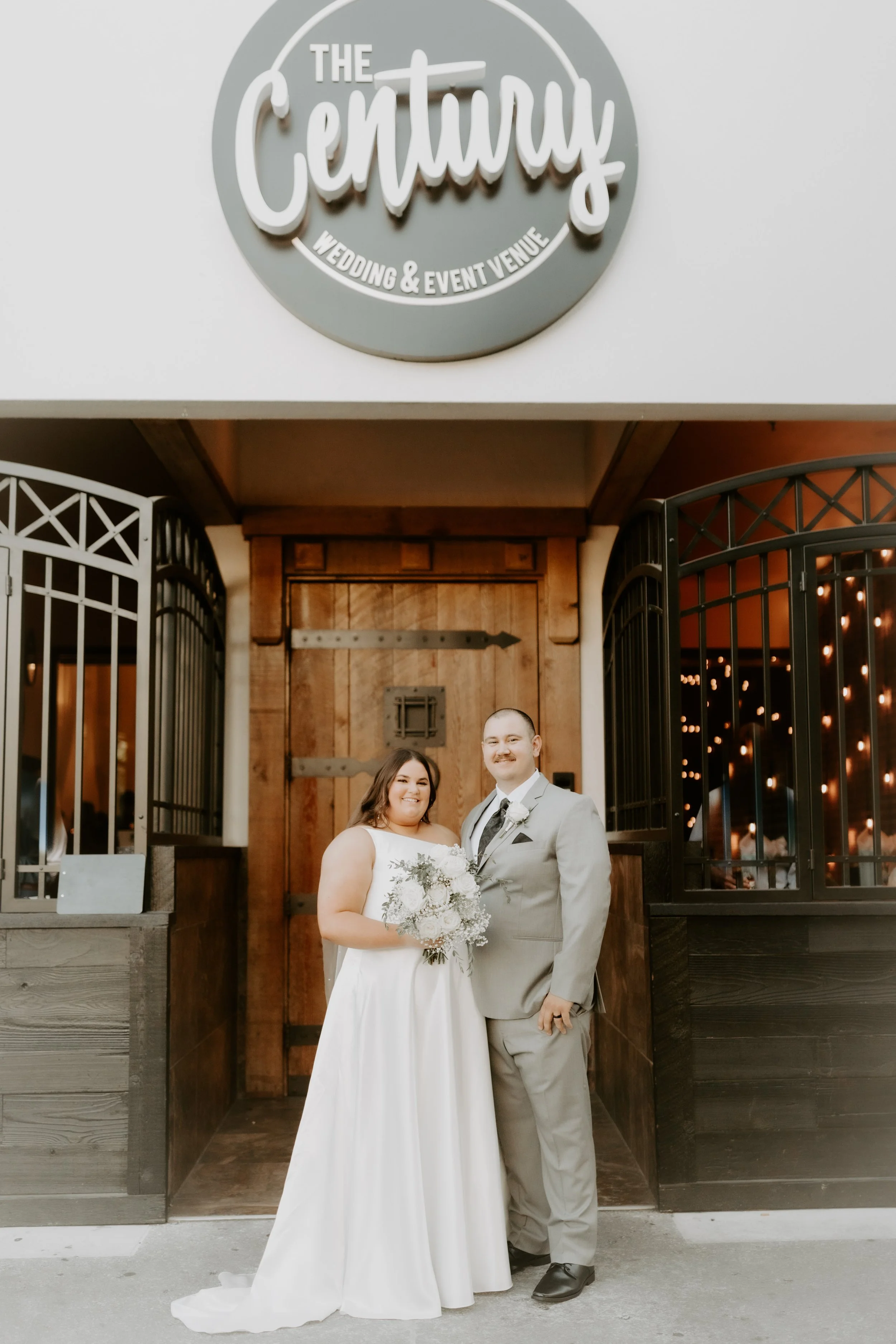 A newlywed couple standing in front of a wedding venue sign that says "The Century Wedding & Event Venue." The bride is holding a bouquet and wearing a white wedding dress, while the groom is dressed in a gray suit. They are smiling and posing for a wedding photo.