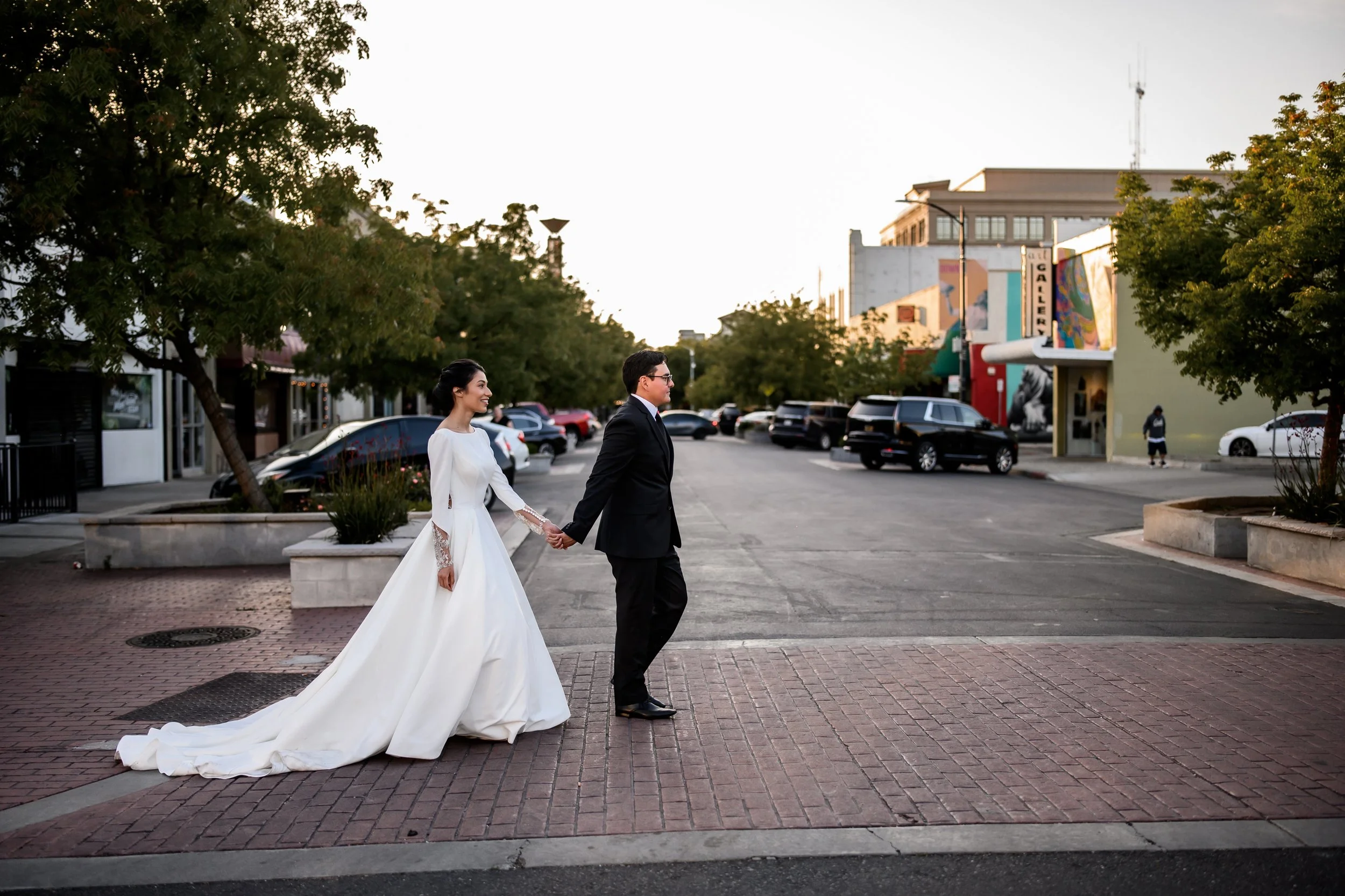 A bride and groom holding hands and walking on a brick sidewalk, with greenery and buildings in the background, during daylight.